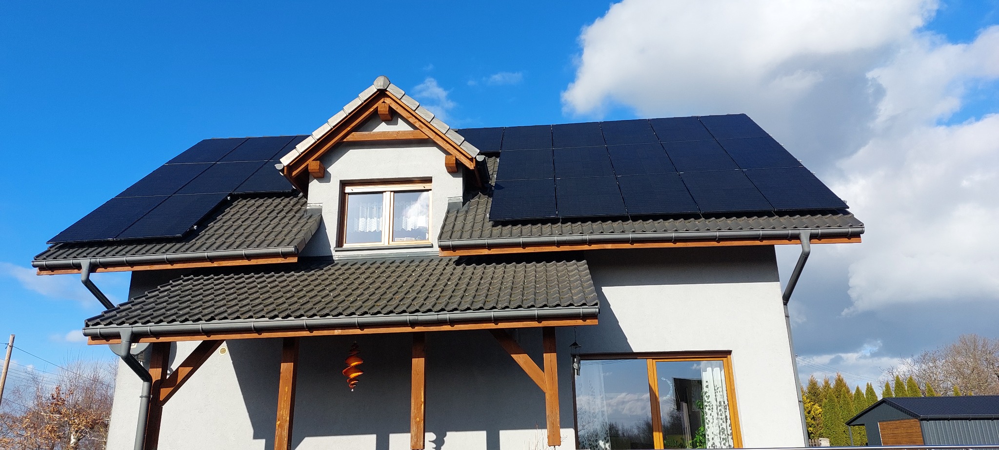 Dom jednorodzinny z panelami fotowoltaicznymi na dachu, widok z dołu na elewację i dach, błękitne niebo z chmurami w tle.