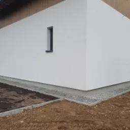 Elewacja budynku z nowo ułożonym obrzeżem z kostki betonowej i podsypką żwirową, widoczne przygotowane podłoże pod trawnik oraz mini koparka w tle.