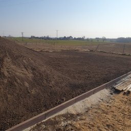AR-KOP ARTUR JAŻDŻEWSKI - Przygotowany teren pod budowę drogi z ułożonymi obrzeżami betonowymi, widoczne świeżo rozplantowana ziemia i elementy drewniane na piasku, w tle pole i zabudowania.