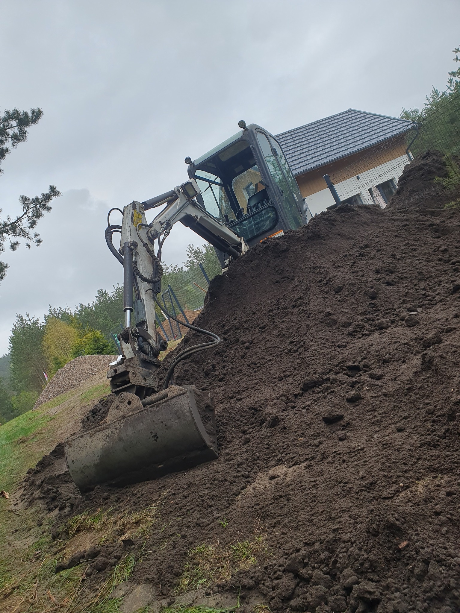 Mała koparka na zboczu usypuje górę ziemi, w tle widoczny dom z szarą dachówką i ogrodzenie z siatki.