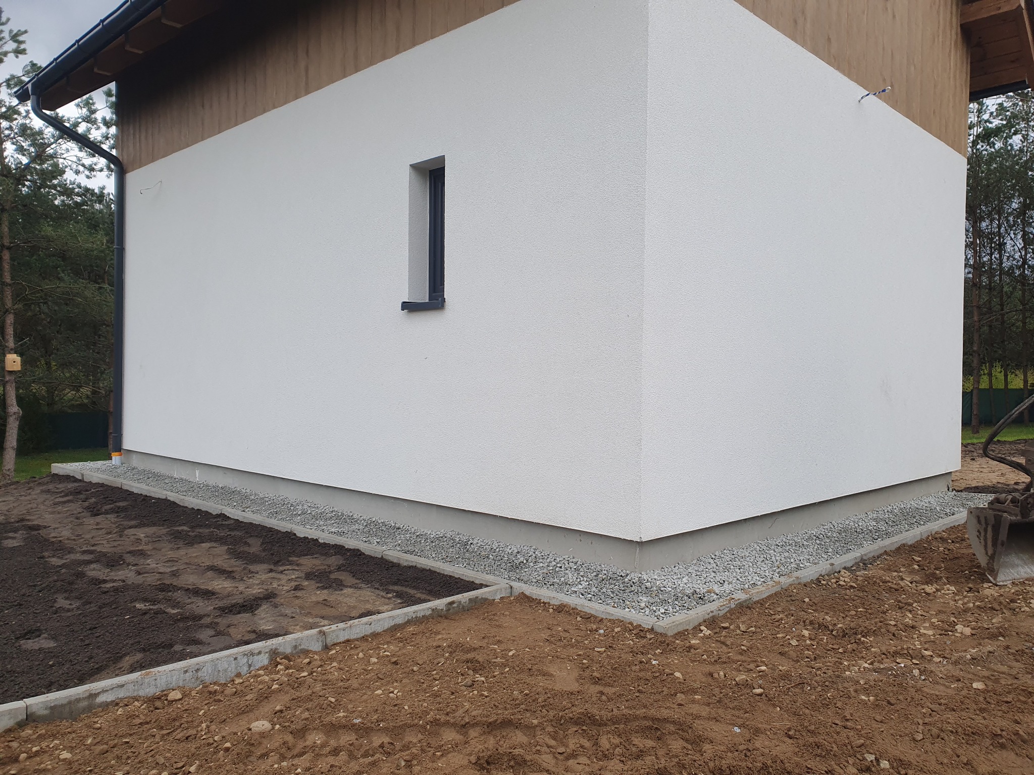 Elewacja budynku z nowo ułożonym obrzeżem z kostki betonowej i podsypką żwirową, widoczne przygotowane podłoże pod trawnik oraz mini koparka w tle.
