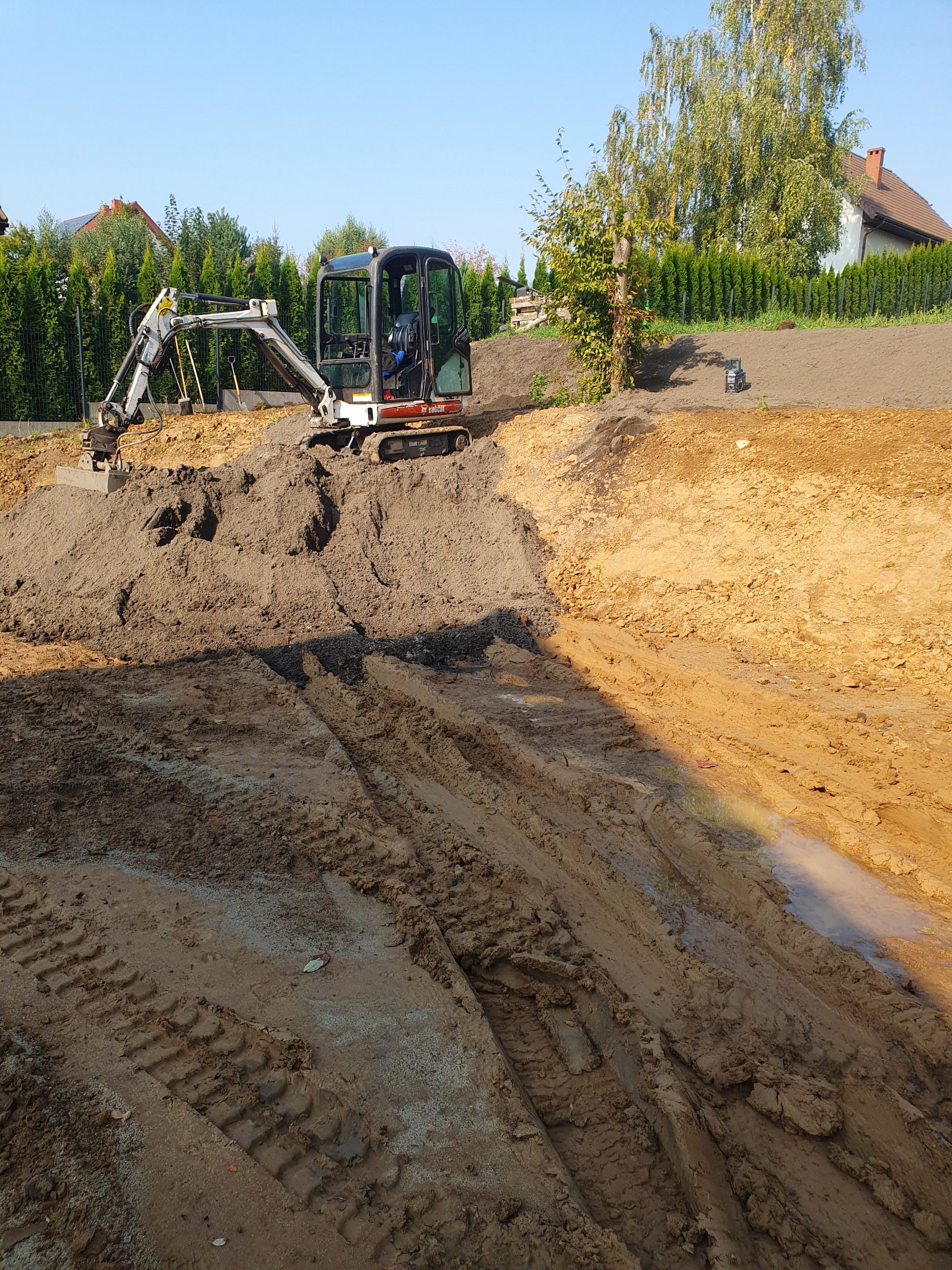 Mała koparka Bobcat na wzniesieniu z ziemi, ślady opon w błocie, przygotowanie terenu pod budowę, widok na domy i drzewa w tle.