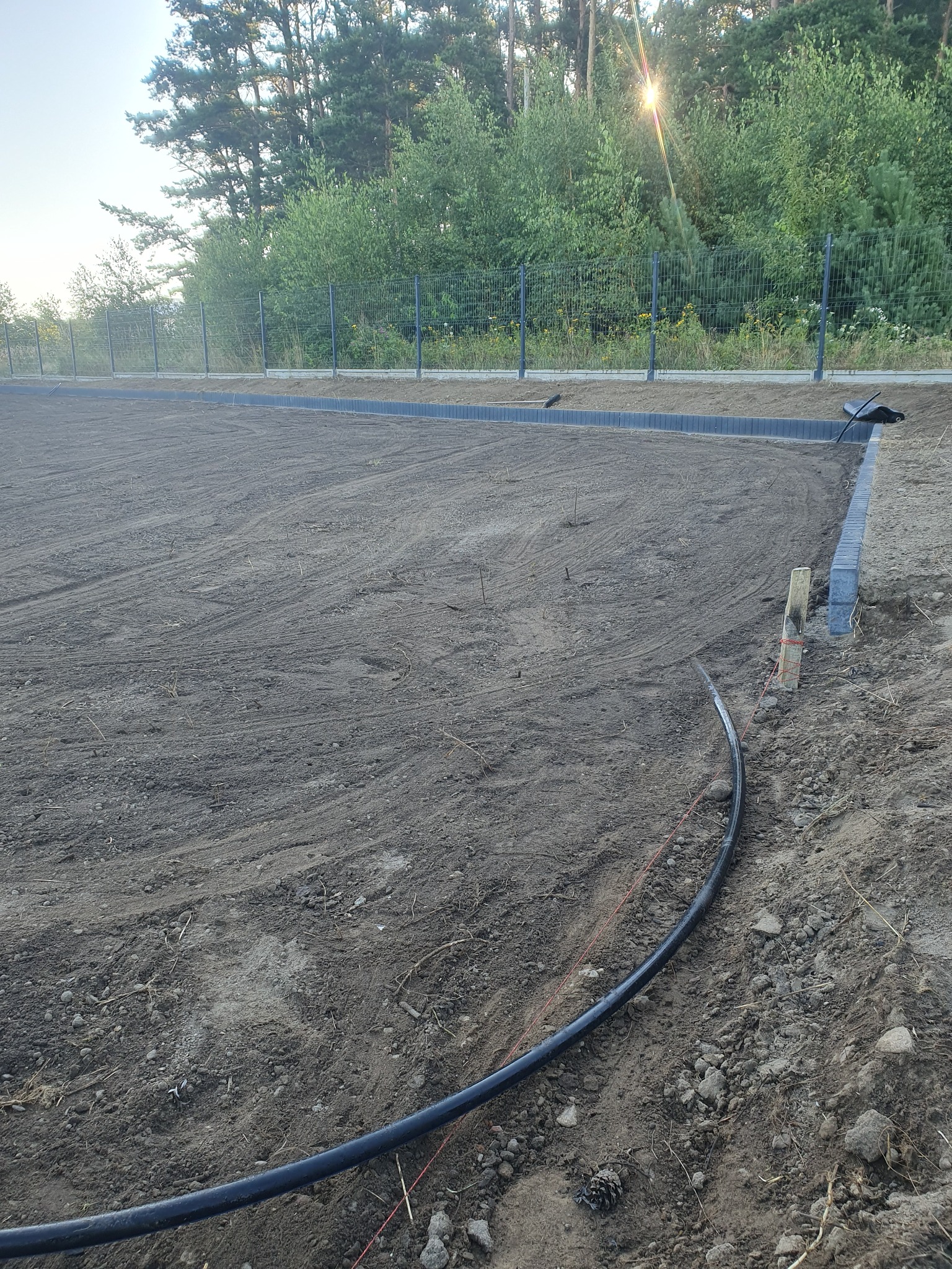 Przygotowany teren pod trawnik z obrzeżami z kostki betonowej i ogrodzeniem panelowym w tle, widoczna rura nawadniająca i wytyczone sznurki.