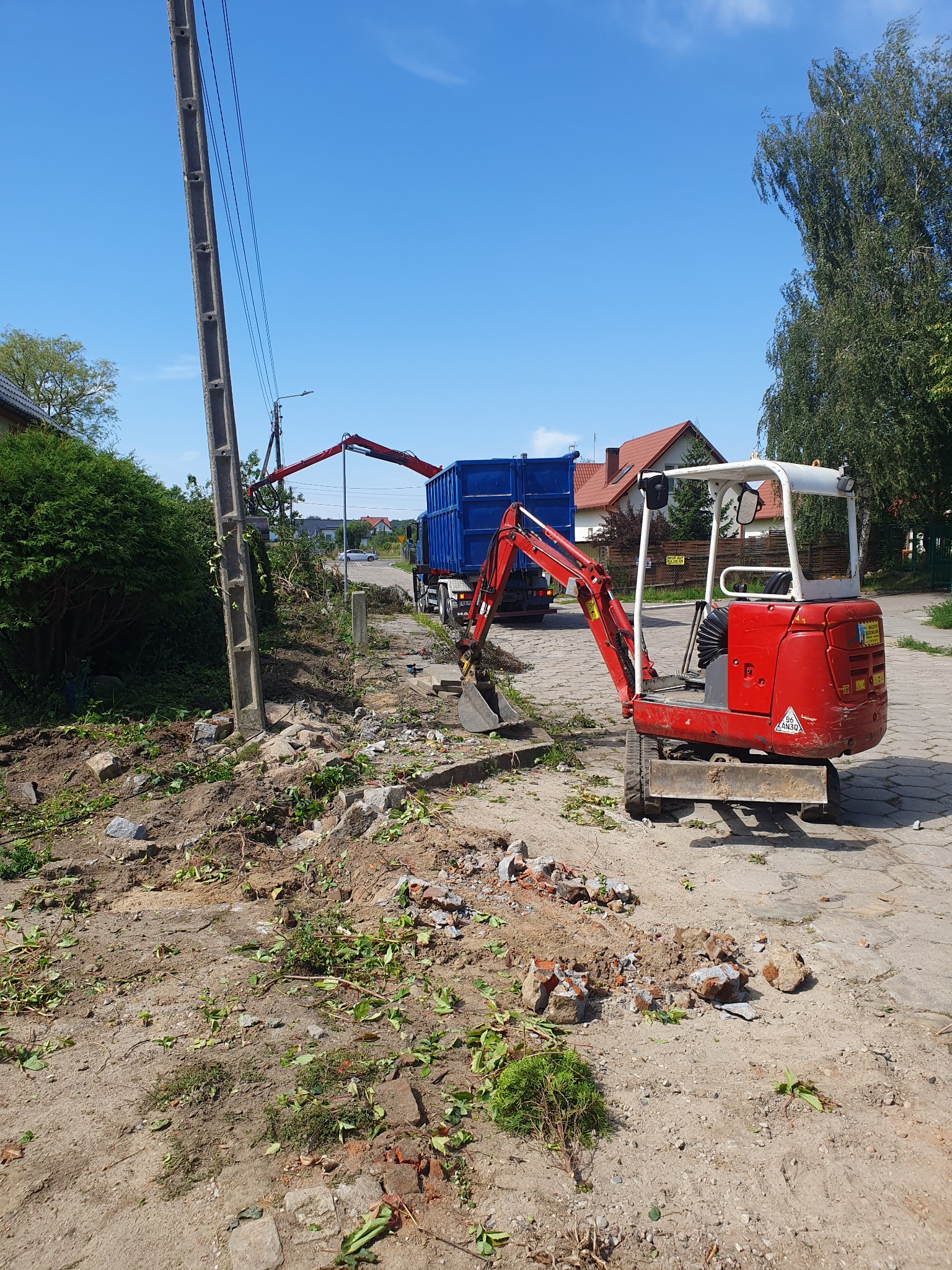 Czerwona mini koparka na tle prac związanych z usuwaniem drzew, z wywrotką i dźwigiem załadowczym w tle, widoczne pozostałości po wycince i gruz.