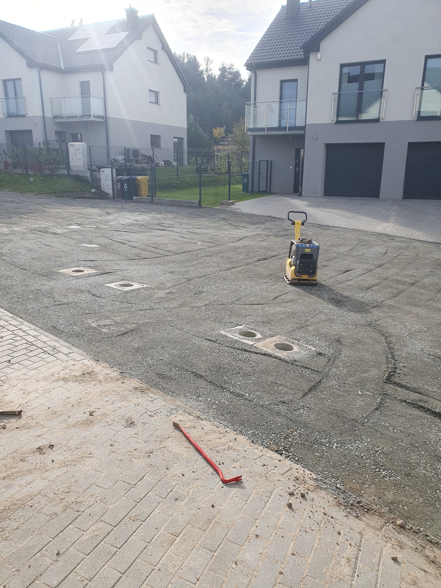 Żółta zagęszczarka wibrująca na żwirowej nawierzchni placu budowy przed nowymi domami, widoczne studzienki i fragment ułożonej kostki brukowej z czerwonym łomem.