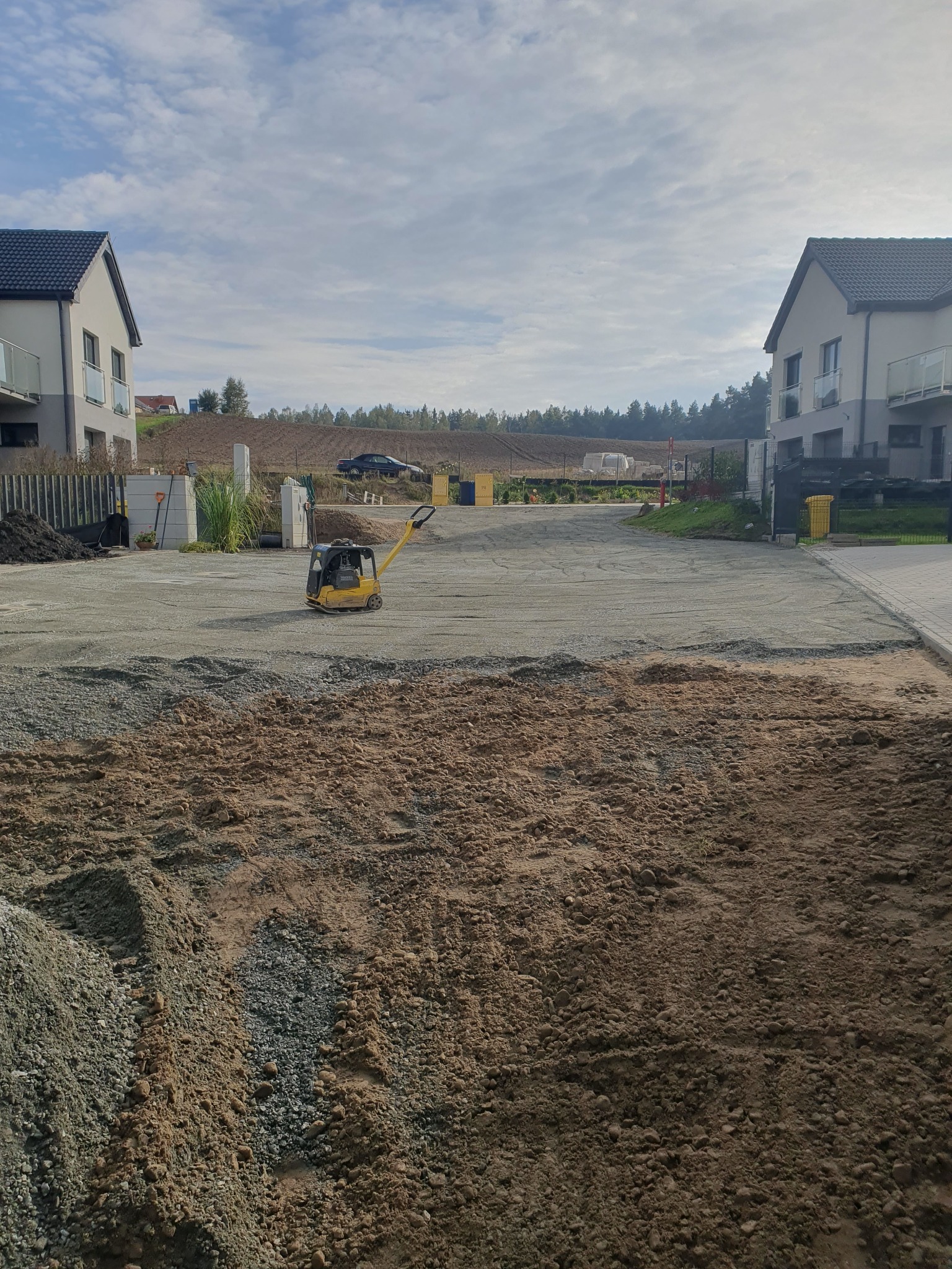 Żółta zagęszczarka wibracyjna Wacker Neuson ubija kamienisty podkład pod drogę dojazdową między dwoma nowymi domami w słoneczny dzień.