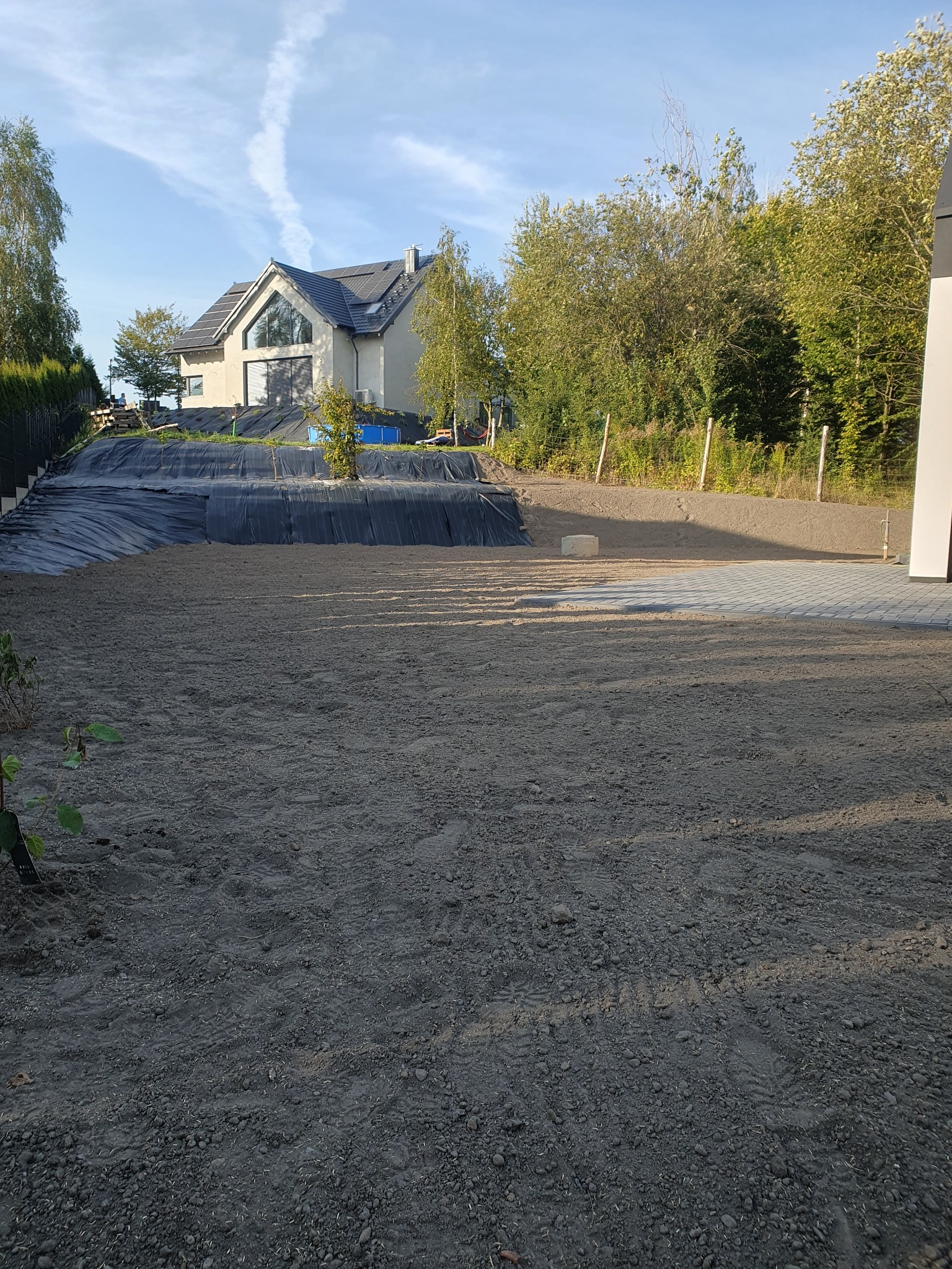 Przygotowany teren pod trawnik, widoczna czarna folia zabezpieczająca skarpę, w tle nowoczesny dom z dużym oknem, częściowo ułożona kostka brukowa.