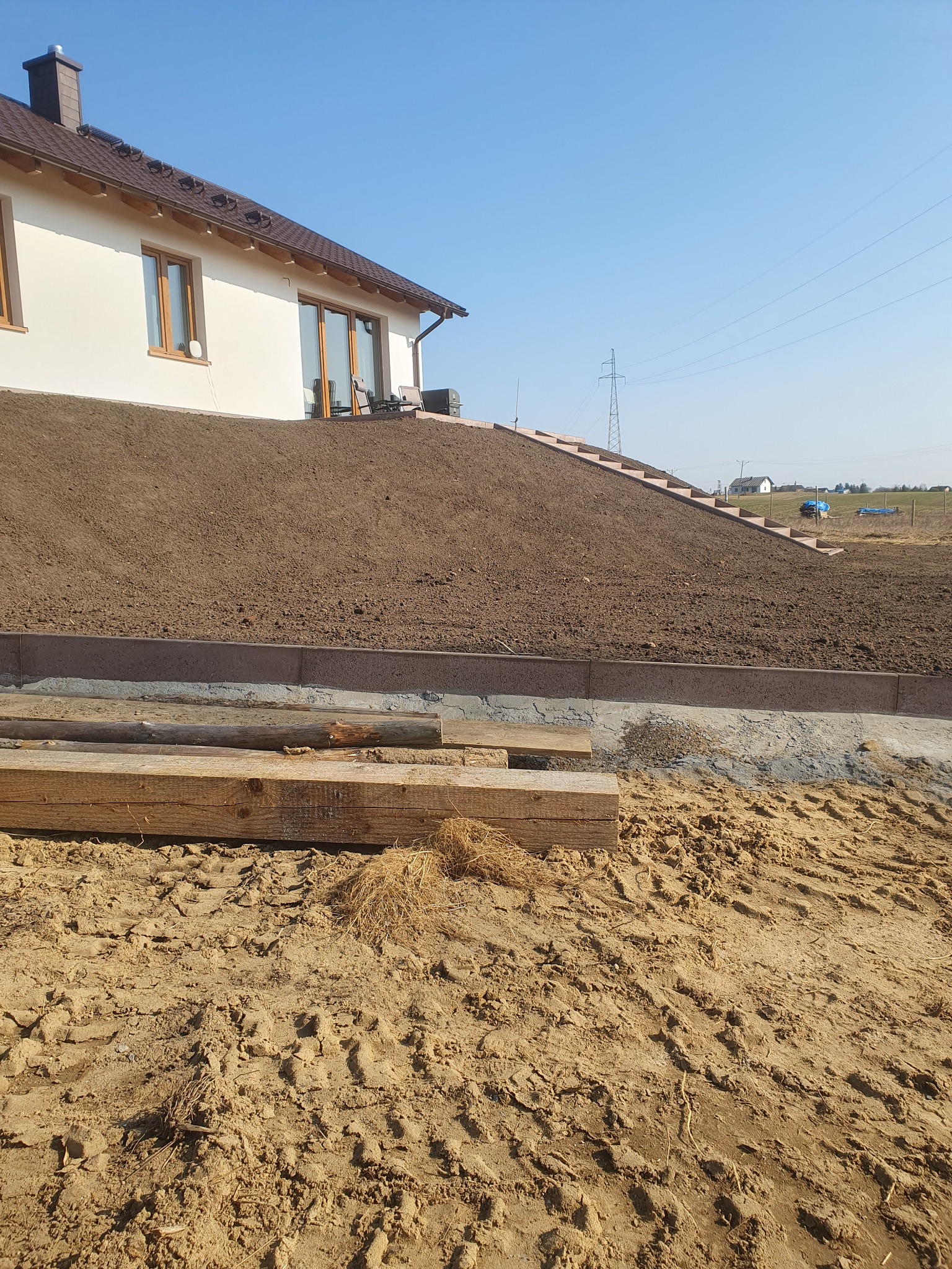 Ukształtowany teren wokół domu, z widocznymi schodami prowadzącymi na górę, zabezpieczony betonową obwódką, w tle dom z brązowym dachem i elementami drewnianymi.