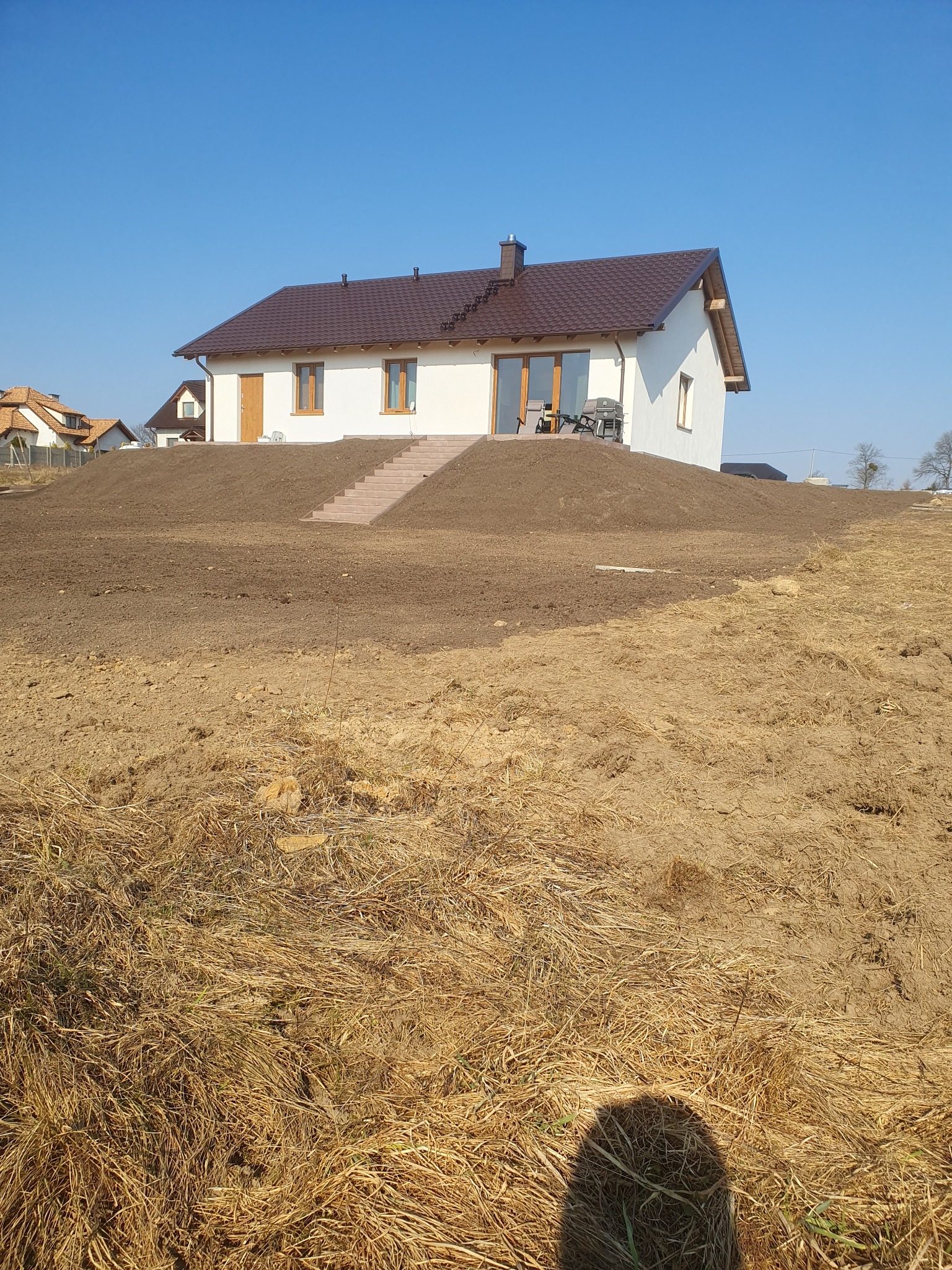 Nowo wybudowany dom z brązowym dachem, stojący na wzgórzu z częściowo przygotowanym terenem pod trawnik, widoczne schody prowadzące do drzwi wejściowych i krzesła na tarasie.