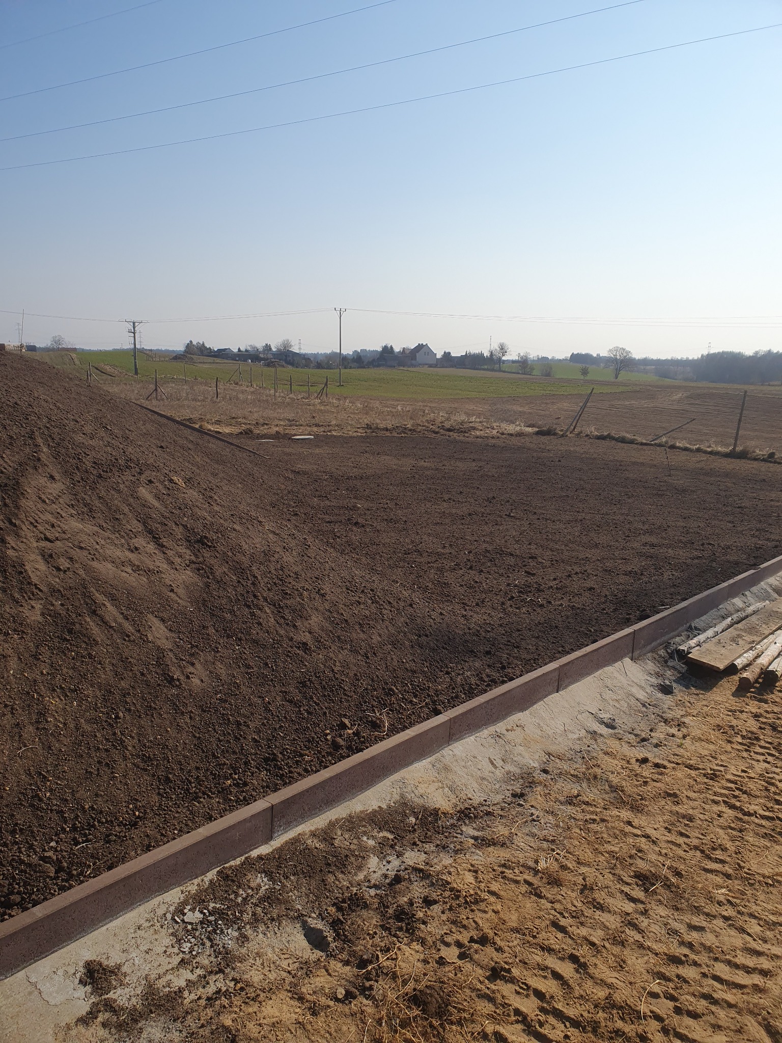Przygotowany teren pod budowę drogi z ułożonymi obrzeżami betonowymi, widoczne świeżo rozplantowana ziemia i elementy drewniane na piasku, w tle pole i zabudowania.