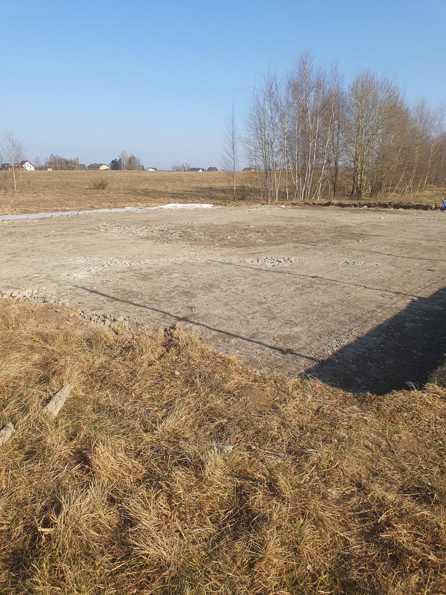 Przygotowany teren pod budowę, wyrównana powierzchnia ziemi z widocznymi śladami po maszynach, otoczona suchą trawą i drzewami bez liści w tle, błękitne niebo.