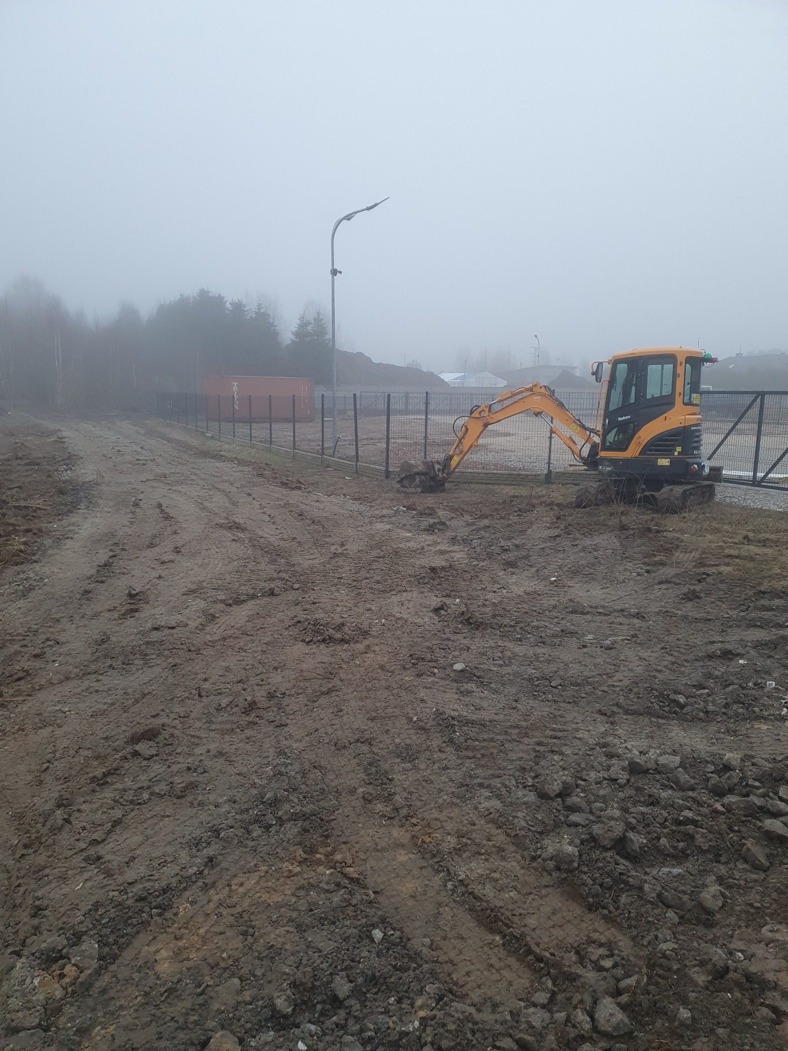 Żółta mini koparka na błotnistym terenie przy ogrodzeniu w mglisty dzień.