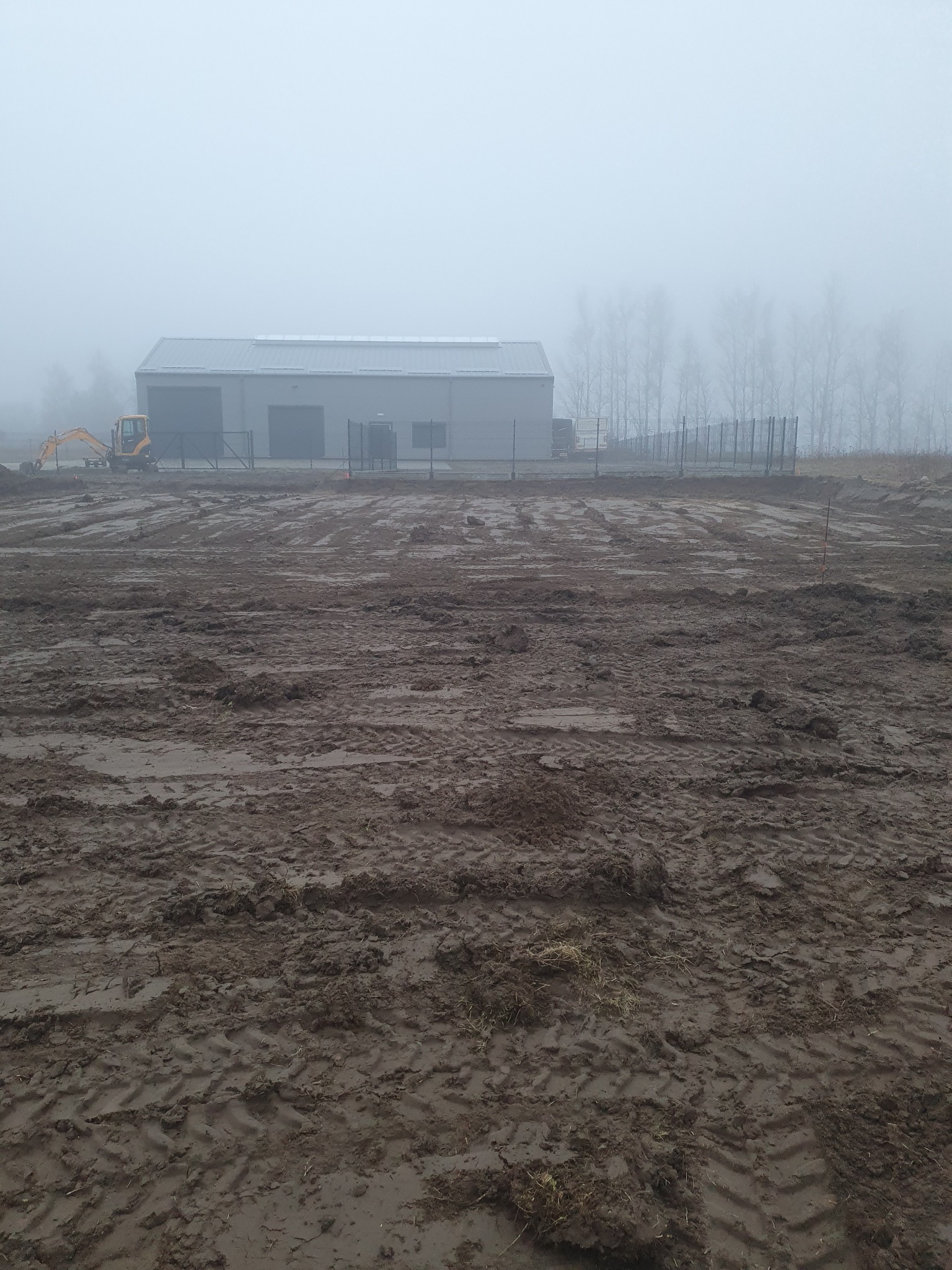 Nierówny, błotnisty teren po niwelacji, widoczny mały żółty koparka, hala magazynowa i ogrodzenie w tle, mglista pogoda.