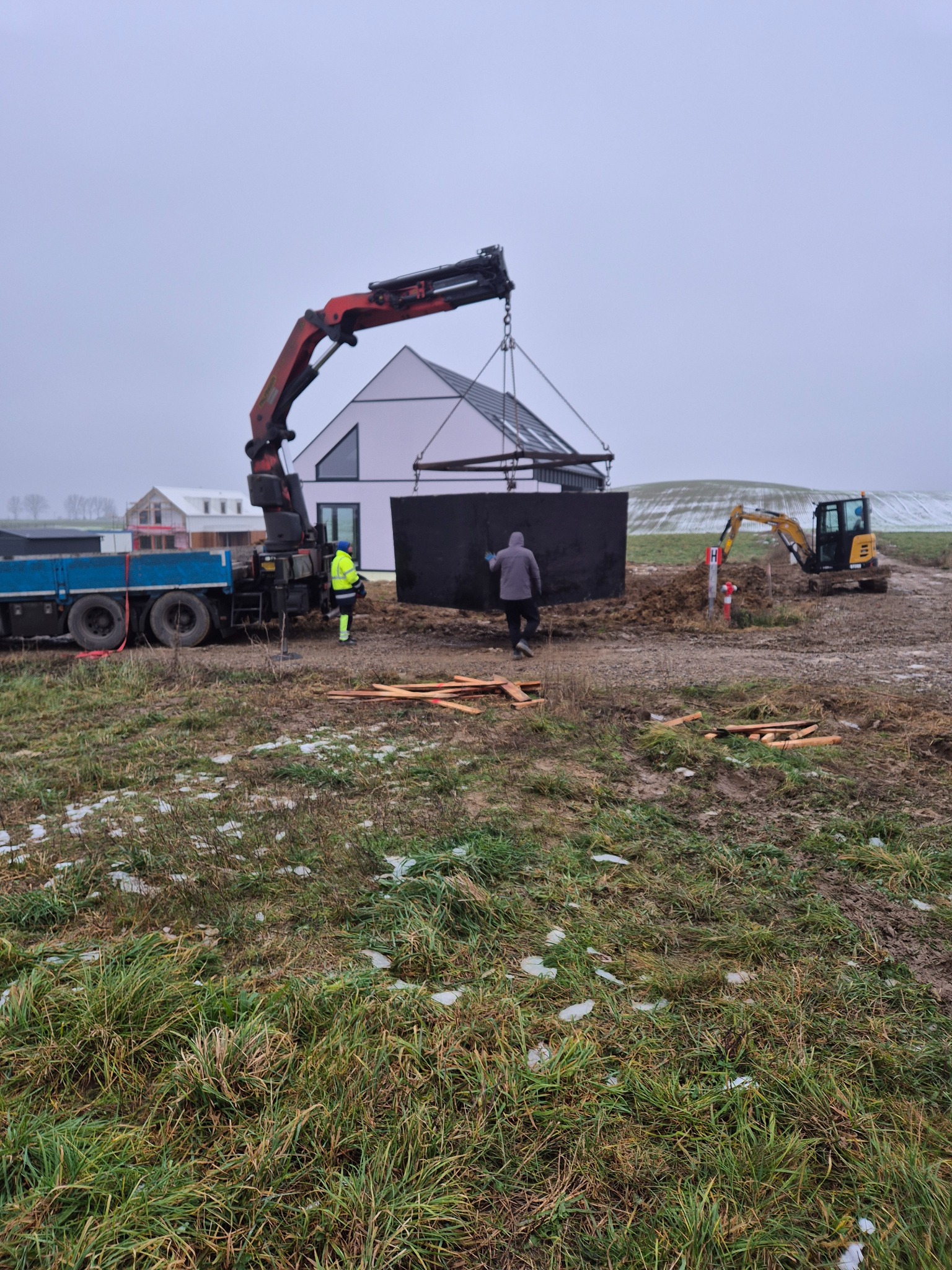 Montaż betonowego szamba za pomocą dźwigu na działce. W tle dom z nowoczesną architekturą i koparka. Pochmurny dzień.