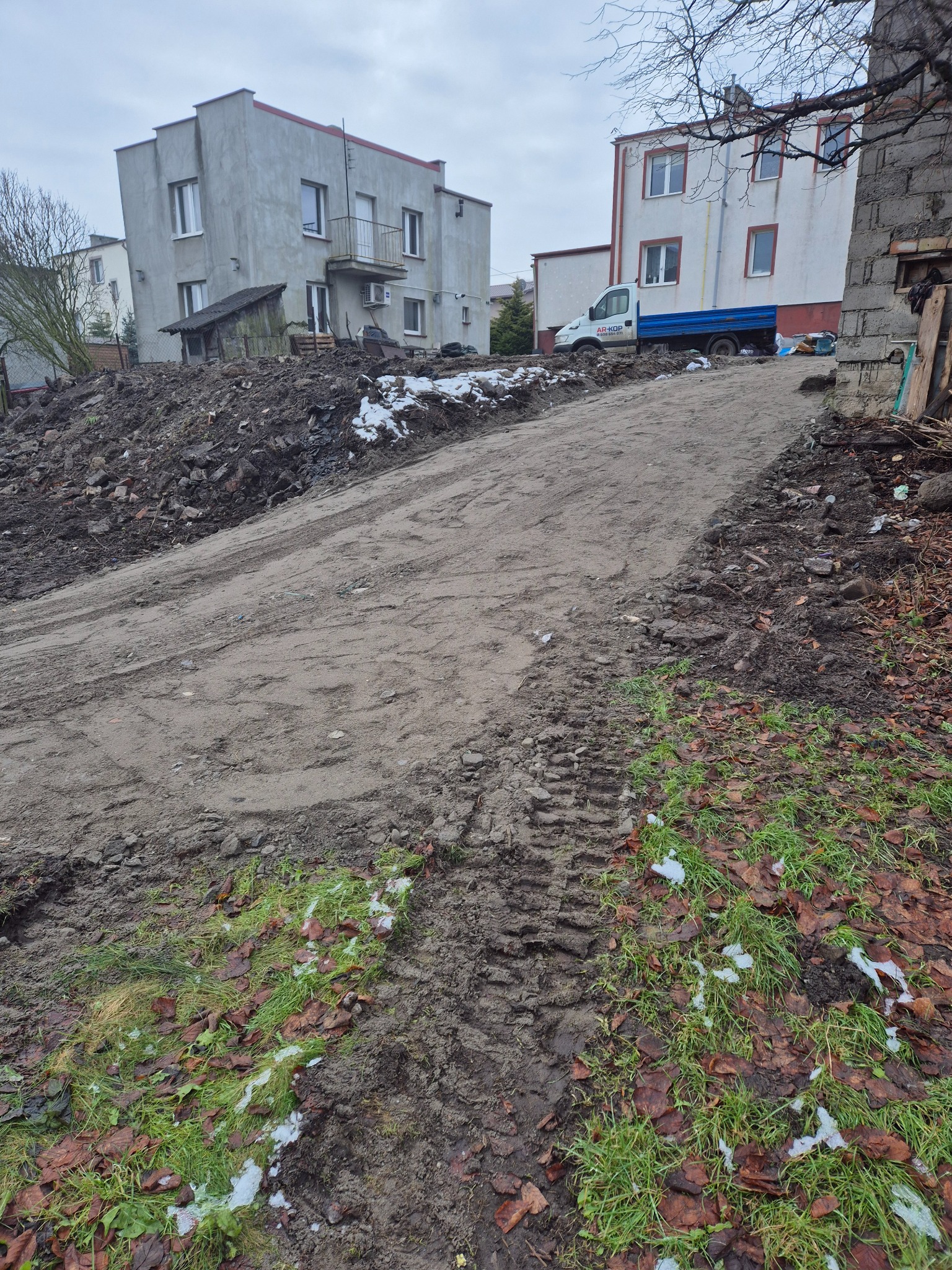 Wyrównany teren pod budowę z widocznymi śladami opon, w tle budynki mieszkalne i samochód dostawczy. Częściowo zachowana roślinność.