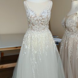 Zakład krawiecki Lussia Olha Mokrytska - Dwa manekiny z sukienkami ślubnymi z koronkowymi zdobieniami. Sukienka na pierwszym planie jest biała, a w tle beżowa.