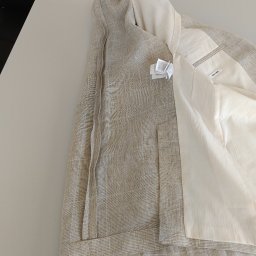 Zakład krawiecki Lussia Olha Mokrytska - Beżowa marynarka w kratę z jasną podszewką, widoczne metki i napis '100% Linen' na wewnętrznej kieszeni, ułożona na płaskiej powierzchni, fragment dłoni w rogu.