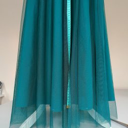 Zakład krawiecki Lussia Olha Mokrytska - Turkusowa, półprzezroczysta tkanina tiulowa mierzona pionowo miarką krawiecką, widoczne białe podbicie i nożyczki na białym stole.