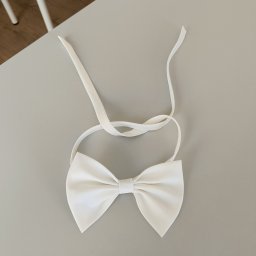 Zakład krawiecki Lussia Olha Mokrytska - Elegancka, biała muszka z satynowego materiału, leżąca na gładkim, szarym stole. Widoczne delikatne załamania materiału i długie paski do zawiązania.