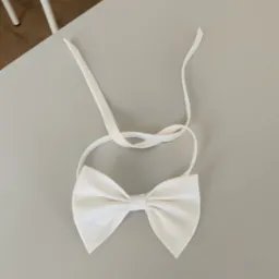 Elegancka, biała muszka z satynowego materiału, leżąca na gładkim, szarym stole. Widoczne delikatne załamania materiału i długie paski do zawiązania.