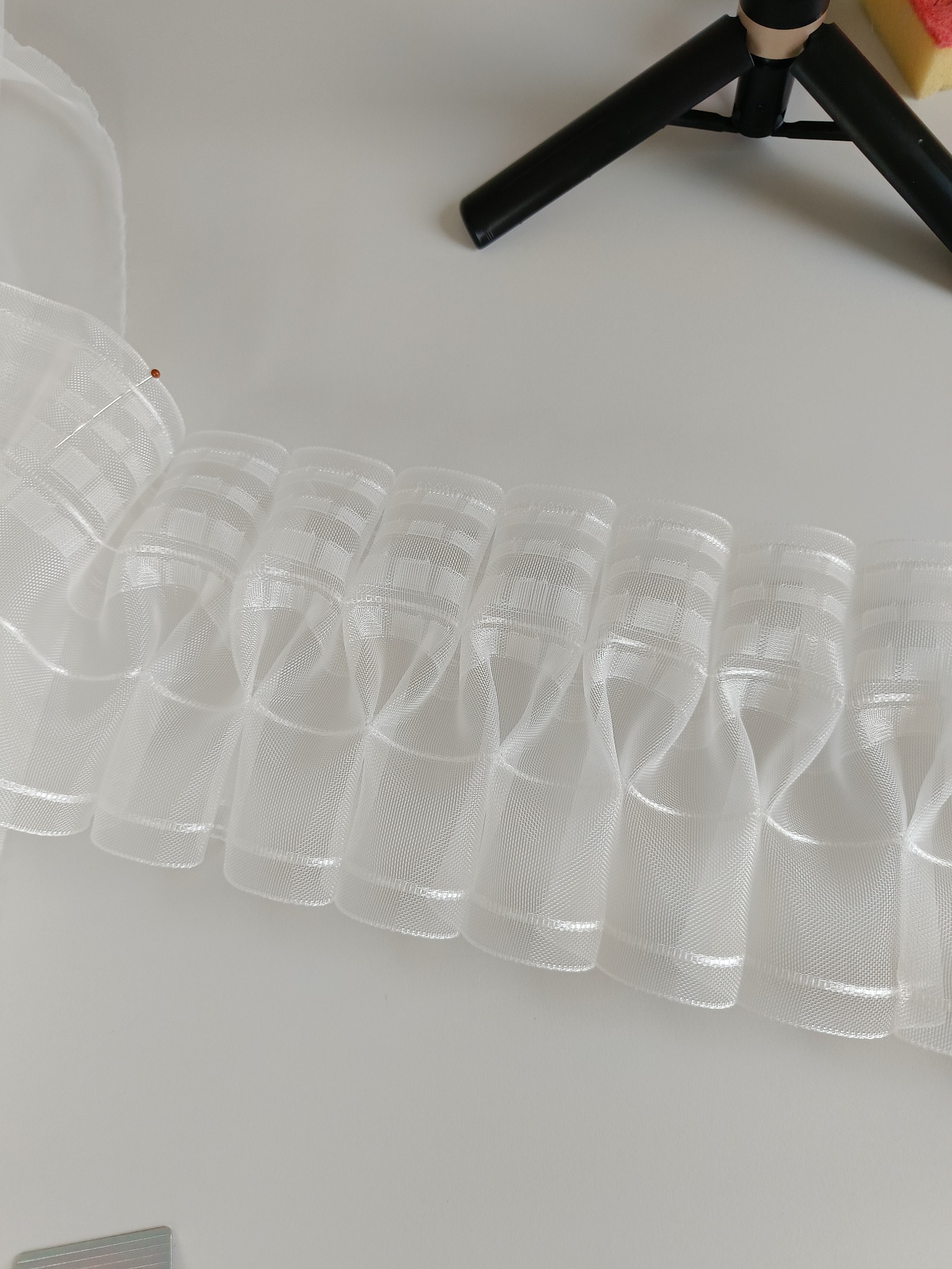 Bliski kadr białej, plisowanej taśmy z delikatnej siateczki z subtelnym wzorem w kratkę, leżącej na białym tle, z widocznym fragmentem czarnego statywu w tle.