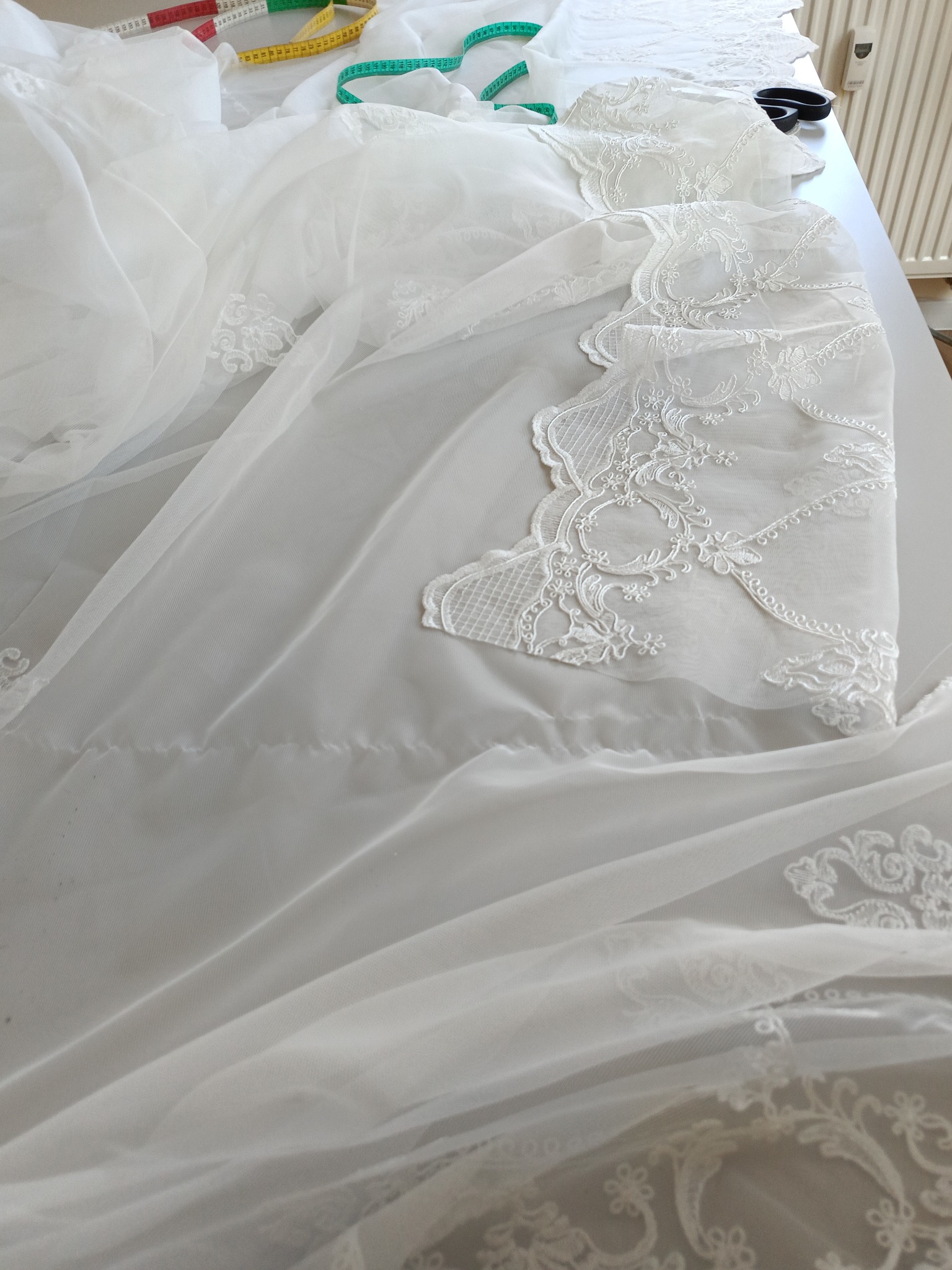 Delikatna, biała tkanina z koronkowymi aplikacjami rozłożona na stole krawieckim, obok leżą miarki krawieckie i nożyczki.