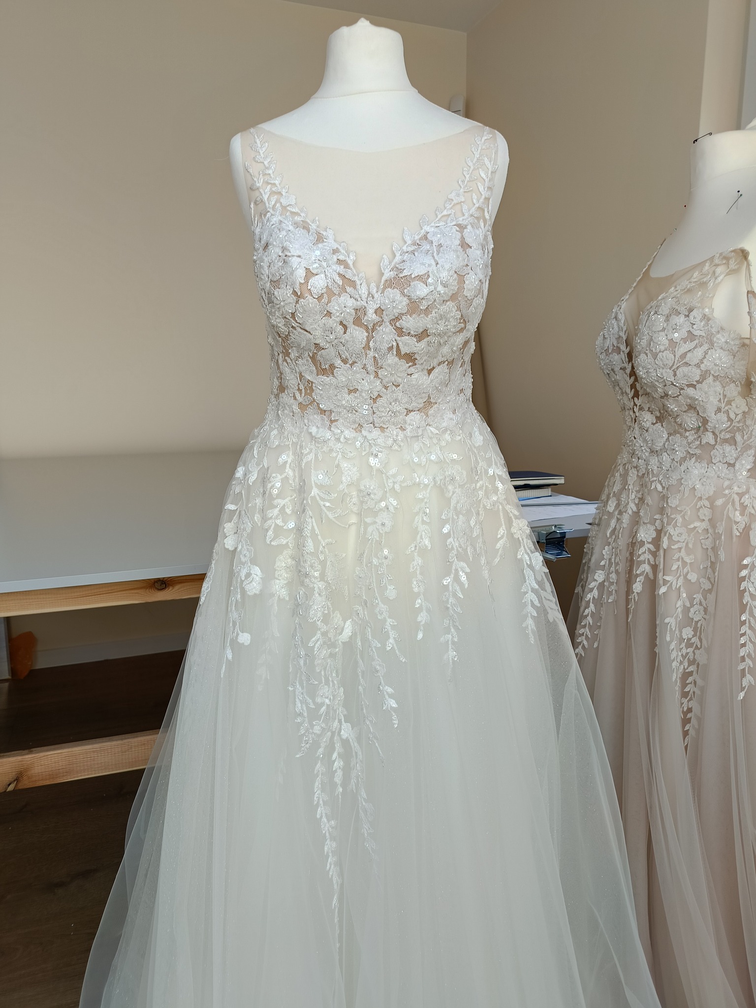 Dwa manekiny z sukienkami ślubnymi z koronkowymi zdobieniami. Sukienka na pierwszym planie jest biała, a w tle beżowa.