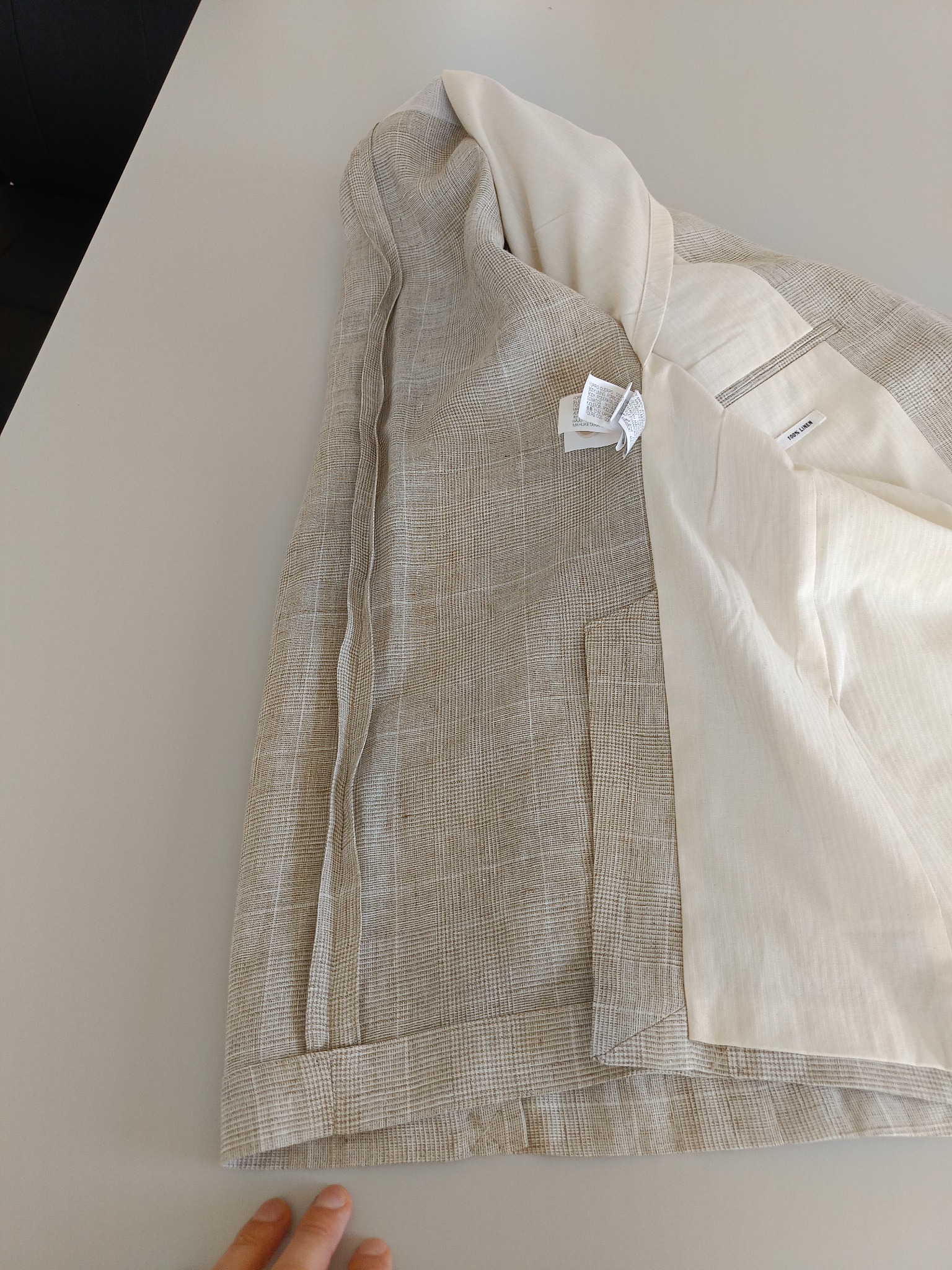 Beżowa marynarka w kratę z jasną podszewką, widoczne metki i napis '100% Linen' na wewnętrznej kieszeni, ułożona na płaskiej powierzchni, fragment dłoni w rogu.