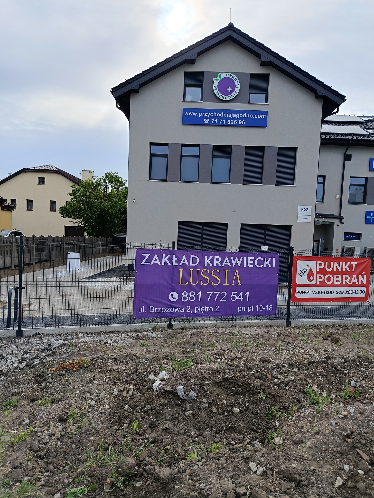 Budynek z szyldem Zakładu Krawieckiego Lussia, widoczny przez ogrodzenie, z ziemią na pierwszym planie.