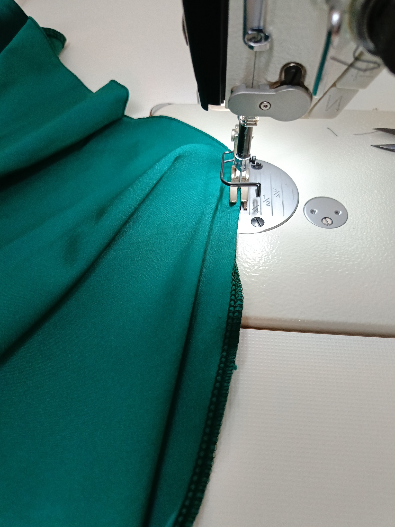Proces szycia na maszynie: szmaragdowy materiał pod igłą maszyny do szycia, widoczny ścieg i miarka na stopce.