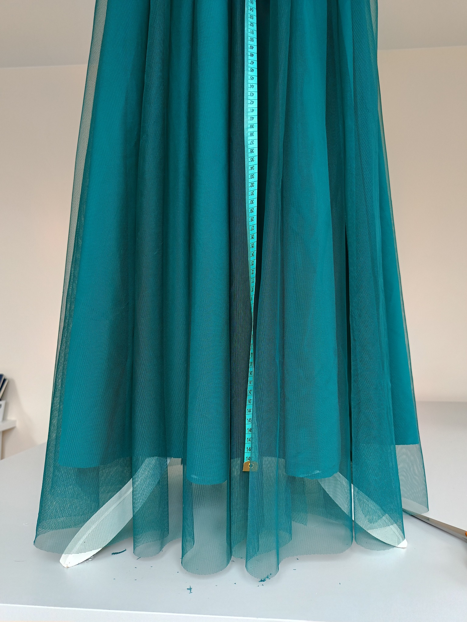 Turkusowa, półprzezroczysta tkanina tiulowa mierzona pionowo miarką krawiecką, widoczne białe podbicie i nożyczki na białym stole.
