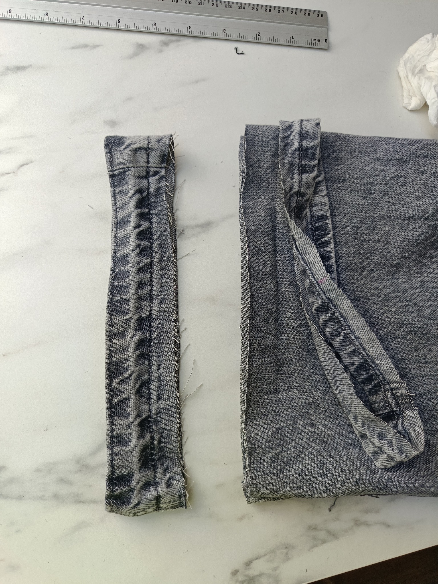 Fragmenty dżinsowej tkaniny, jeden wąski pasek z postrzępionymi brzegami, drugi większy kawałek złożony z podwiniętym brzegiem, leżące na białym marmurowym blacie obok metalowej linijki.