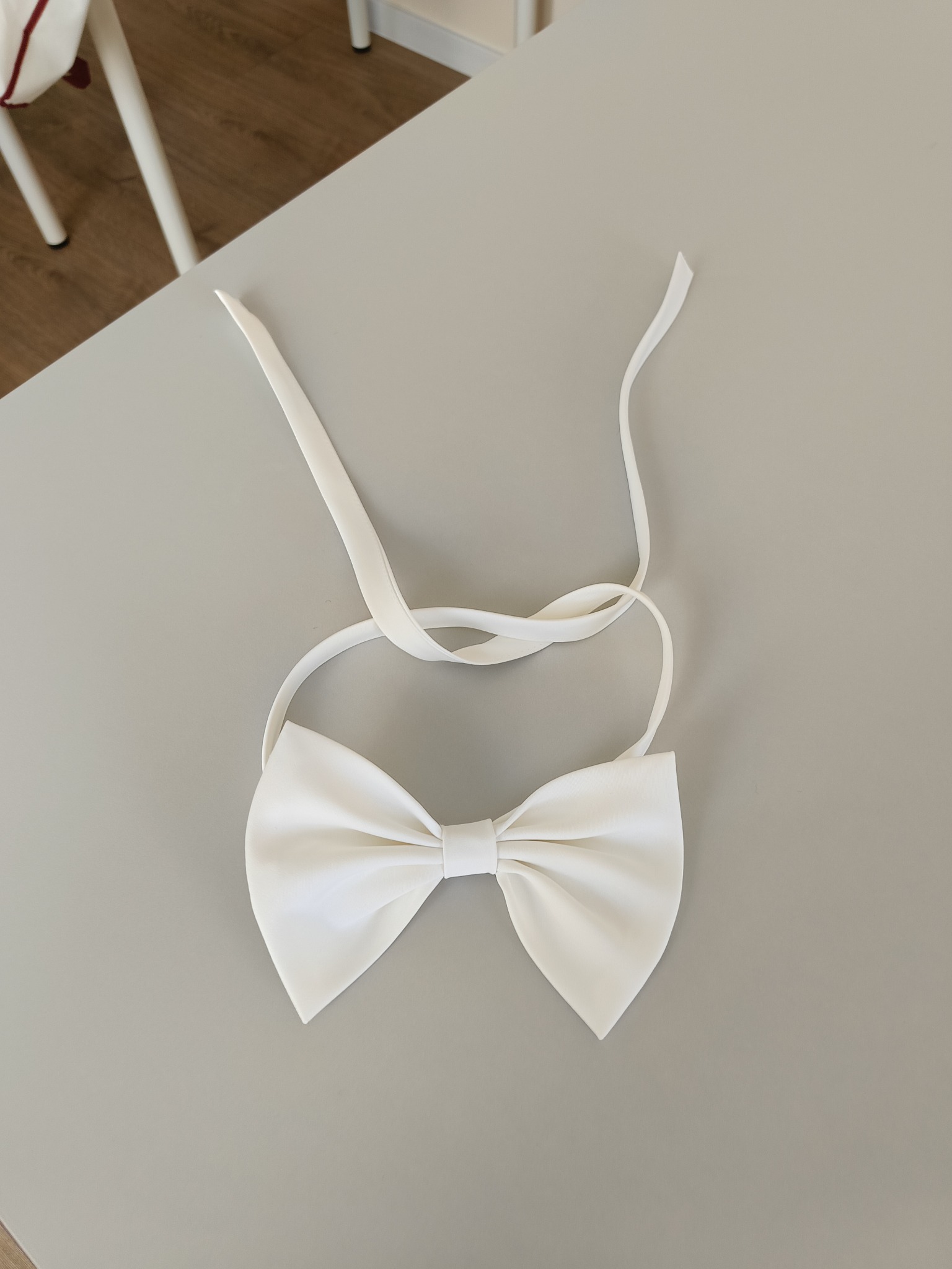 Elegancka, biała muszka z satynowego materiału, leżąca na gładkim, szarym stole. Widoczne delikatne załamania materiału i długie paski do zawiązania.