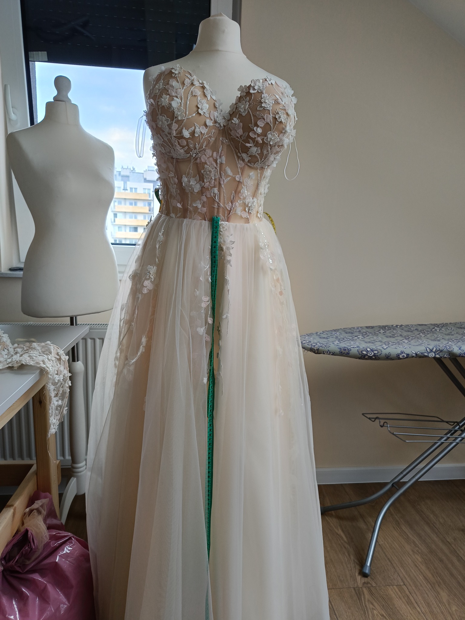 Suknia ślubna z delikatnymi kwiatowymi aplikacjami na manekinie krawieckim, w tle widoczne okno i drugi manekin, obok deska do prasowania