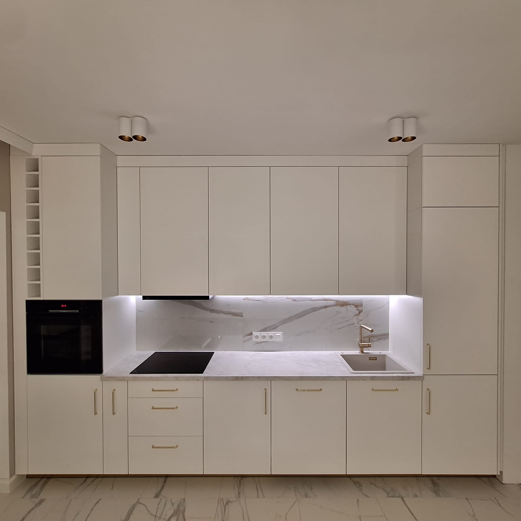 Nowoczesna, jasna kuchnia z białymi frontami szafek, złotymi uchwytami i blatem imitującym marmur. Widoczna płyta indukcyjna, piekarnik i złota bateria przy zlewie. Nad blatem podświetlana ściana...