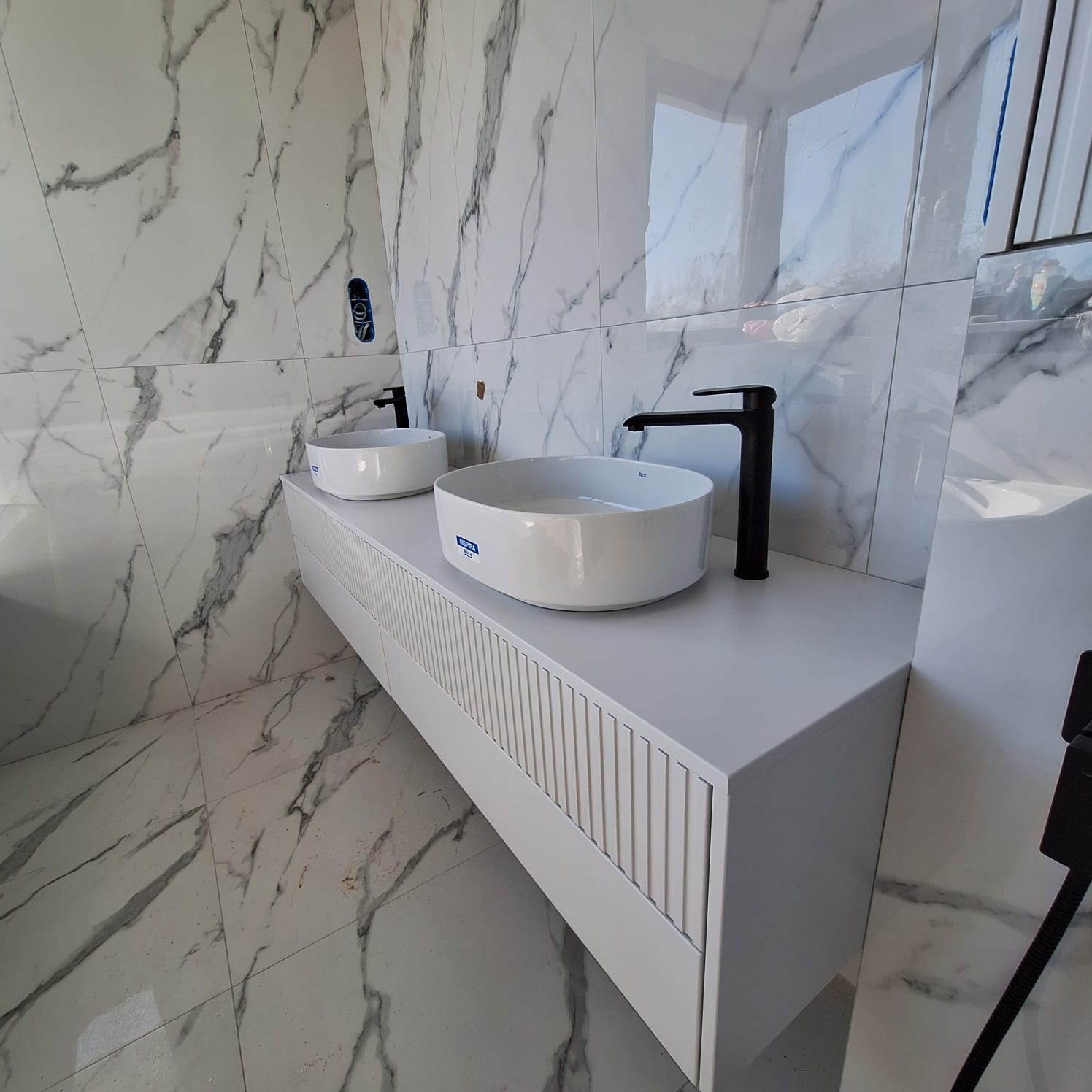 Nowoczesna łazienka z dwoma okrągłymi umywalkami nablatowymi, czarną armaturą i białą szafką z frezowanymi frontami na tle marmurowych płytek.