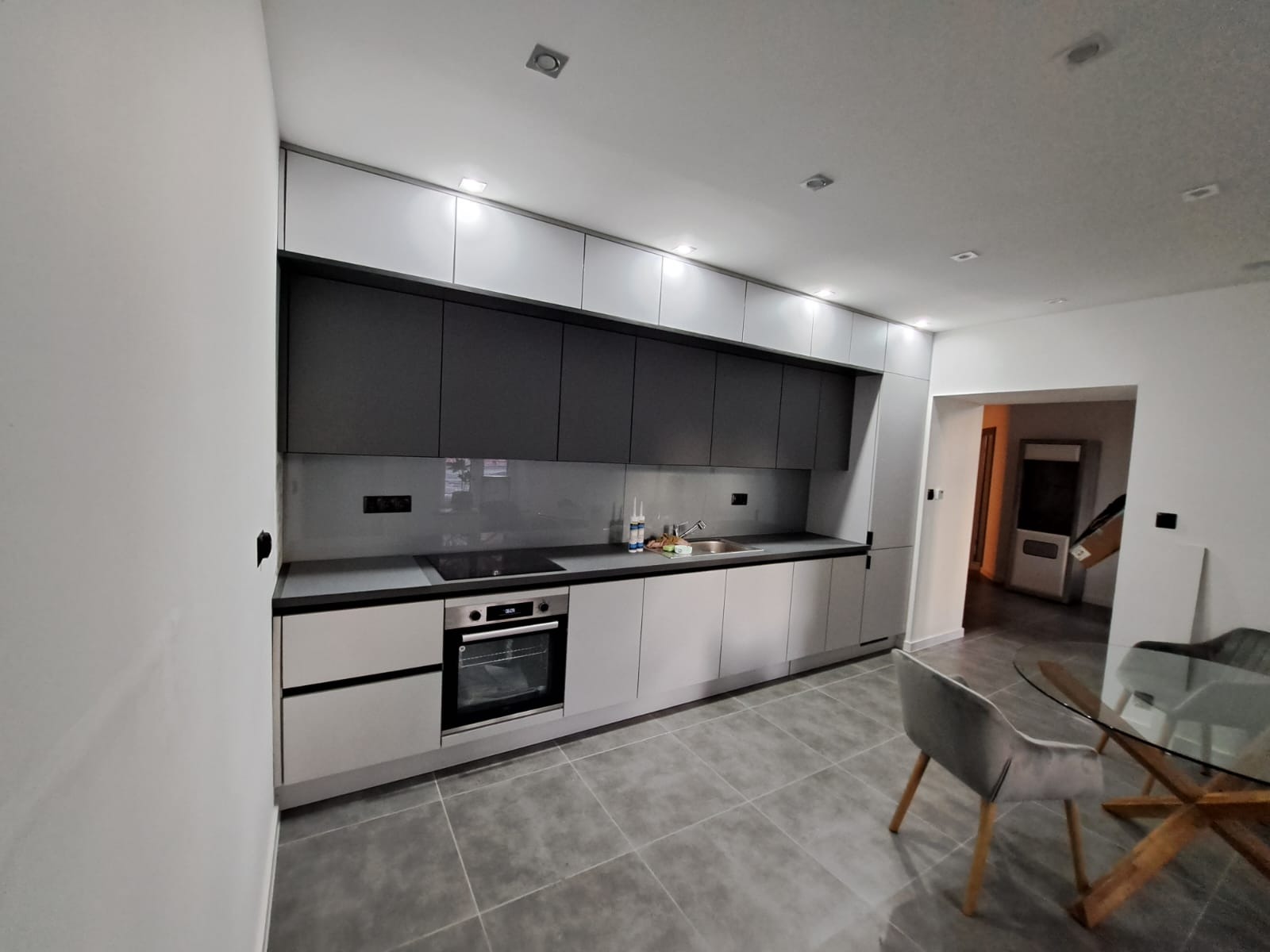 Nowoczesna kuchnia z szarymi szafkami górnymi i jasnoszarymi dolnymi, szklany panel nad blatem, płyta indukcyjna i piekarnik w zabudowie, widok na jadalnię z okrągłym szklanym stołem.