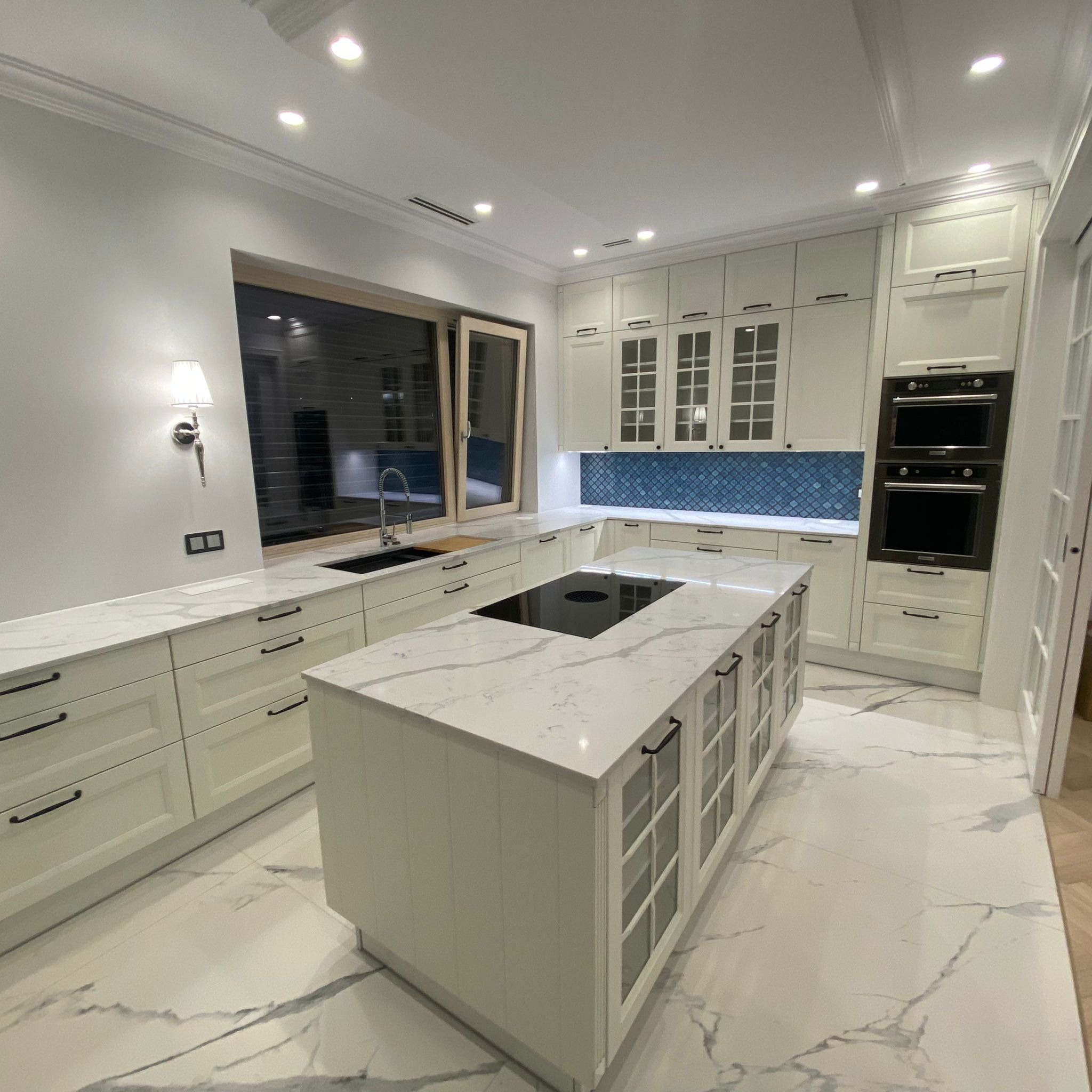 Elegancka, jasna kuchnia z wyspą, białymi frontami szafek z czarnymi uchwytami, marmurową podłogą i niebieską mozaiką nad blatem roboczym. Oświetlenie punktowe i dekoracyjny kinkiet.