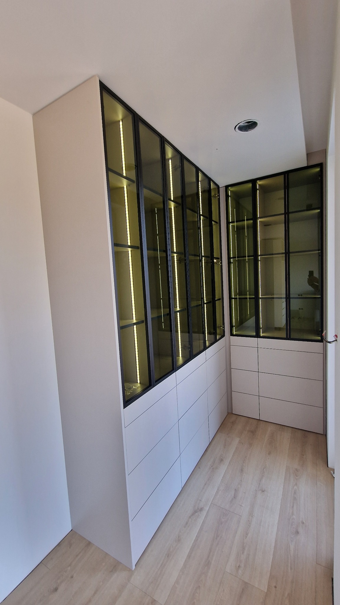 Nowoczesna szafa wnękowa z podświetlanymi półkami za szklanymi drzwiami, zintegrowana z komodą z szufladami, na podłodze panele imitujące jasne drewno.