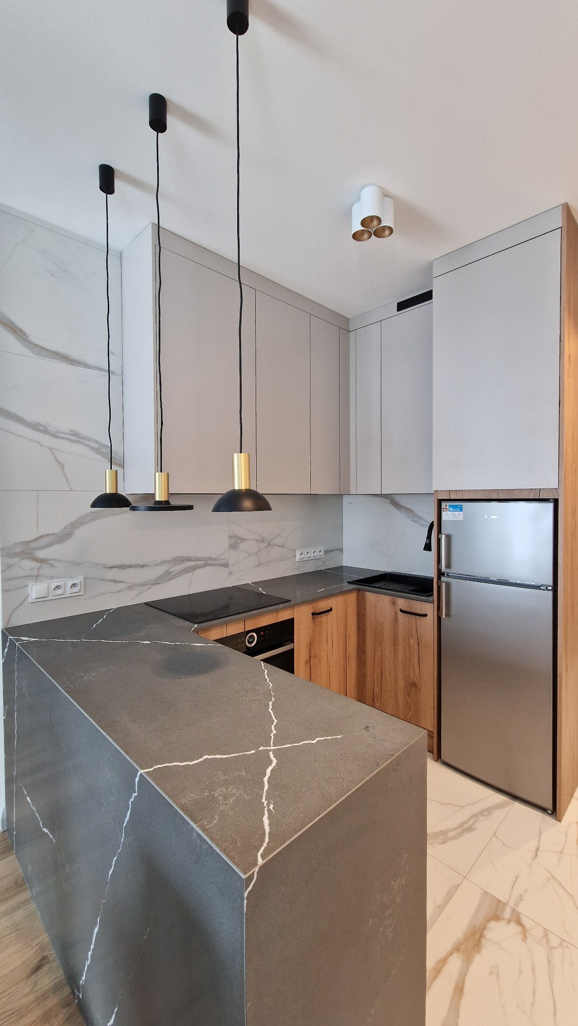 Nowoczesna kuchnia z wyspą, szare szafki, drewnopodobne fronty, marmurowe płytki na ścianie i podłodze, czarne lampy wiszące nad wyspą, srebrna lodówka.