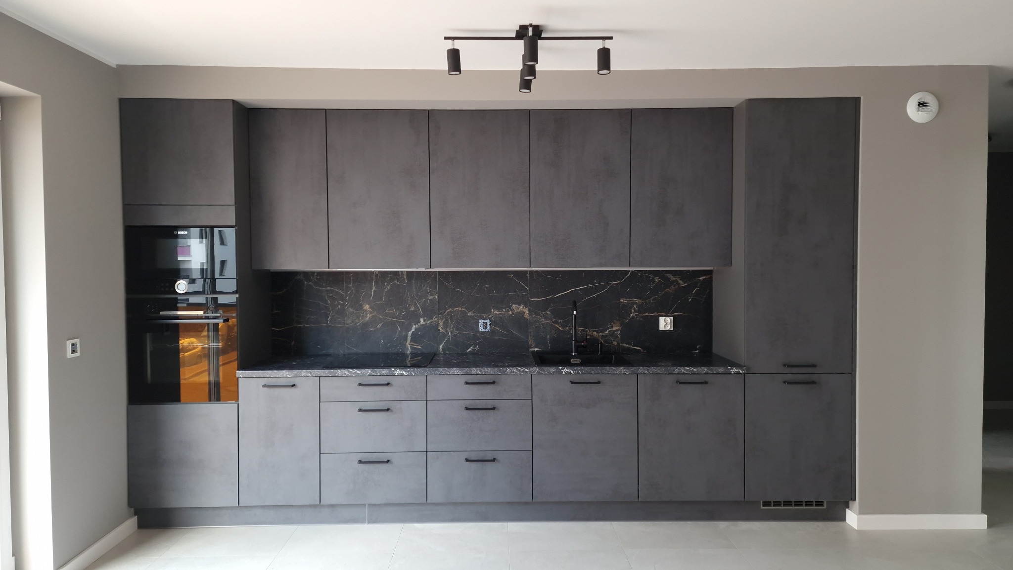 Nowoczesna kuchnia z szarymi matowymi frontami, czarnym blatem i marmurowym backsplashiem, oświetlona czarnymi reflektorami sufitowymi.