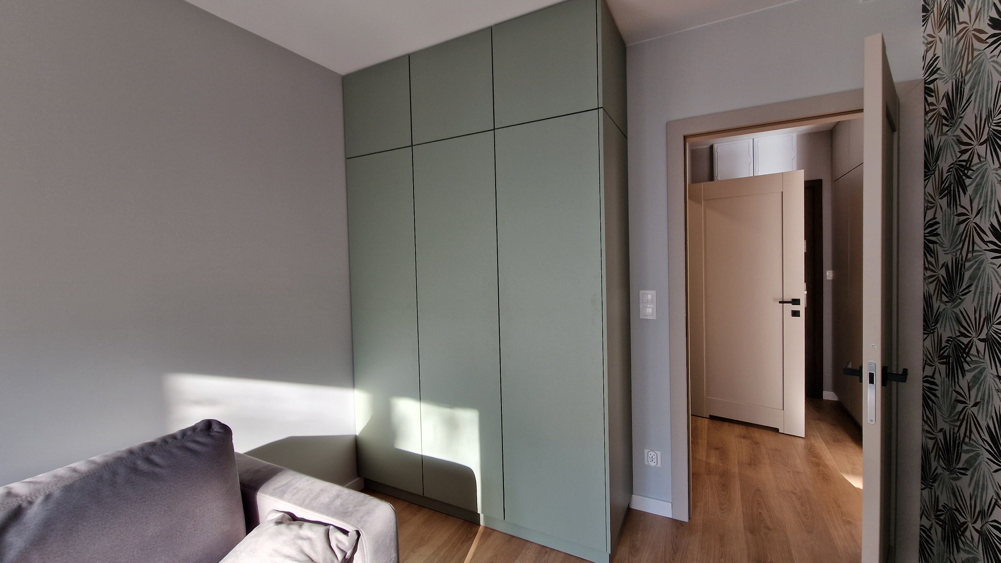 Pistacjowa szafa z frontami bez uchwytów, z widocznym salonem w tle i otwartymi drzwiami do kolejnego pomieszczenia, na podłodze panele.