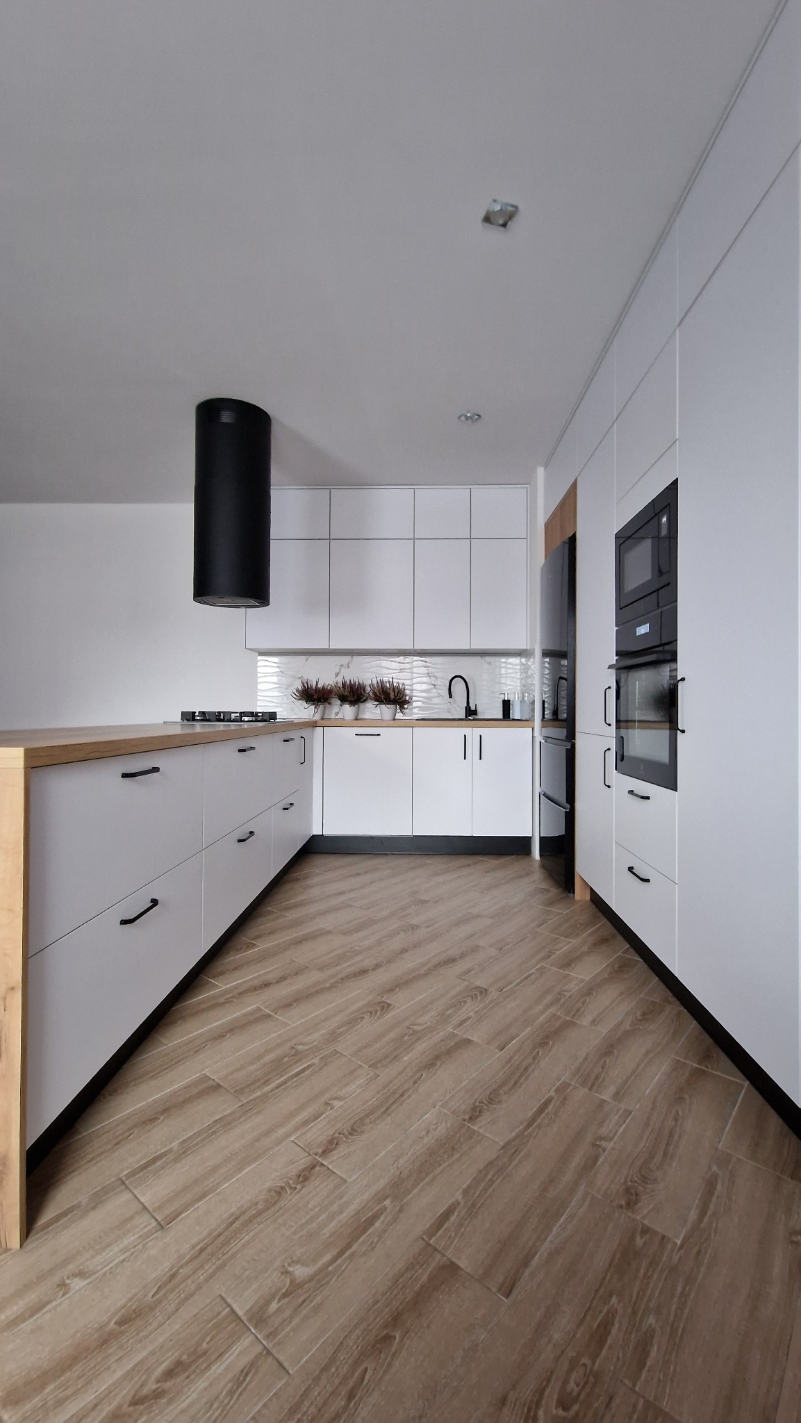 Nowoczesna kuchnia z białymi frontami, drewnianym blatem wyspowym, czarnym okapem cylindrycznym i płytkami imitującymi drewno na podłodze.