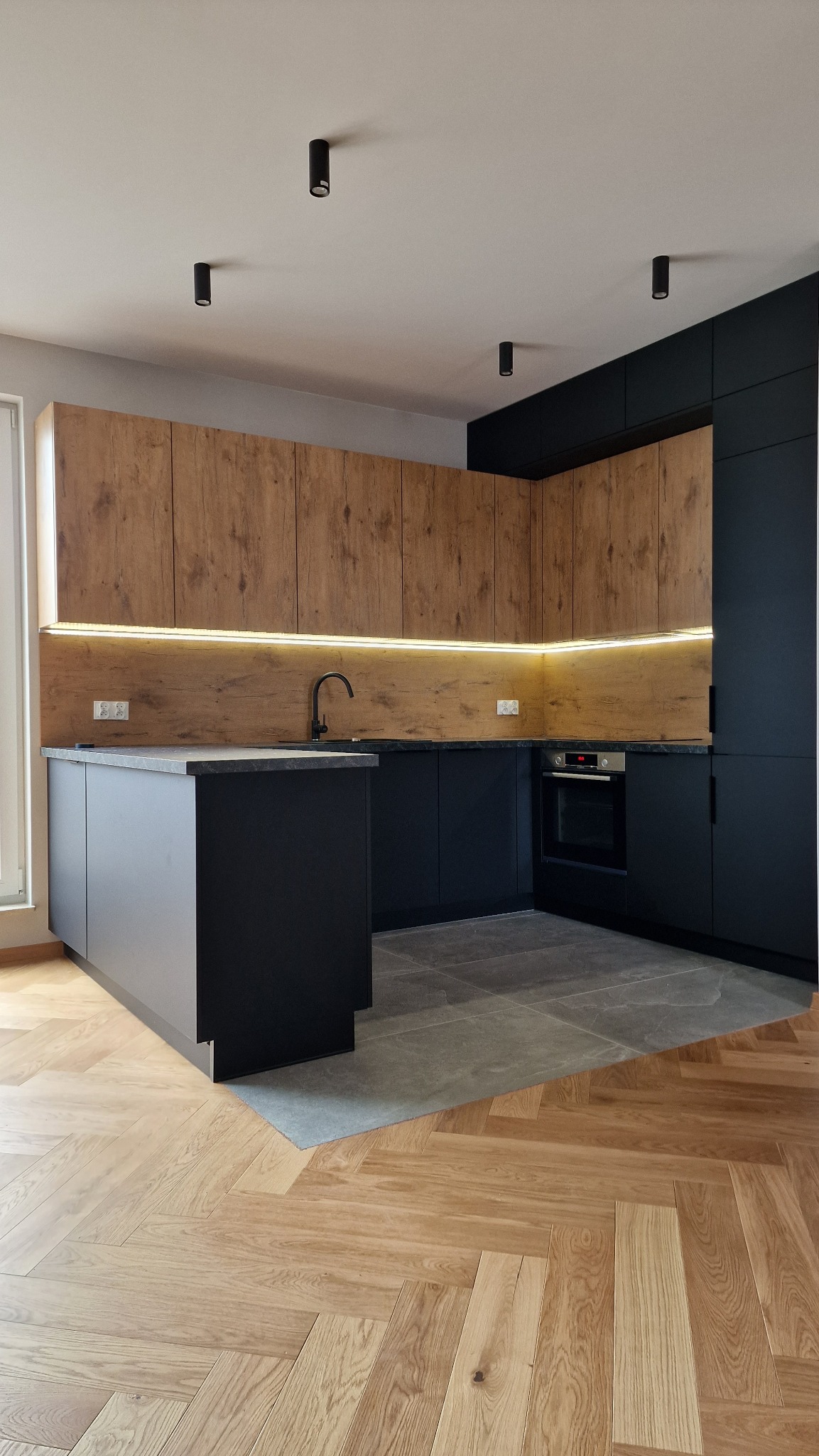 Nowoczesna kuchnia w Bielawie z czarnymi frontami dolnych szafek i drewnopodobnymi górnymi, podświetlonymi LED, czarny zlew i armatura, na podłodze parkiet w jodełkę przechodzący w szare płytki.