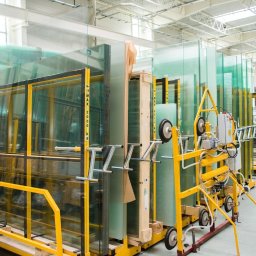Domiplast Warszawa - Szyby szklane ułożone w metalowych, żółtych stojakach w hali produkcyjnej, z widocznym urządzeniem do transportu szyb na pierwszym planie.