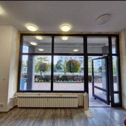 Domiplast Warszawa - Wnętrze pomieszczenia z dużą, przeszkloną ścianą z czarnymi profilami, otwartymi szklanymi drzwiami, grzejnikiem pod oknem i widokiem na ulicę z zielenią.