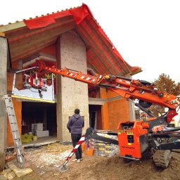 Domiplast Warszawa - Montaż okna z wykorzystaniem specjalistycznego dźwigu przyssawkowego SPX424 na budowie domu jednorodzinnego w stanie surowym otwartym, widoczny pracownik nadzorujący proces.