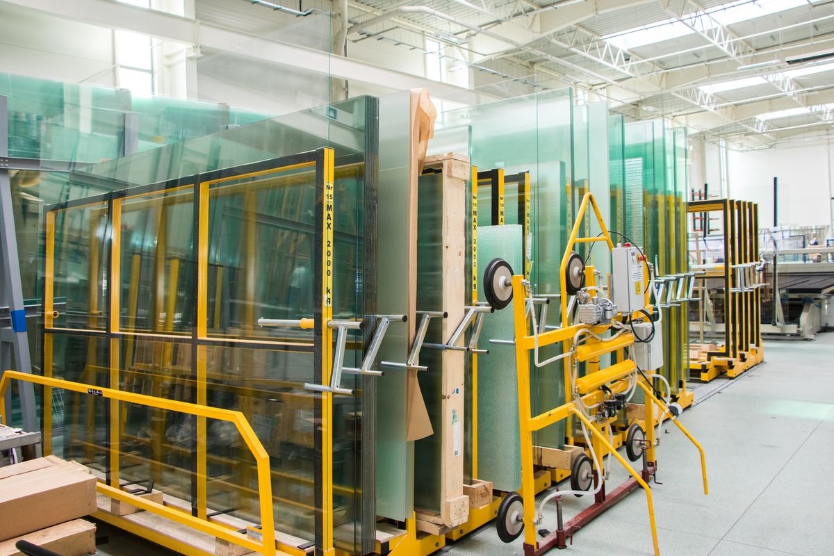 Szyby szklane ułożone w metalowych, żółtych stojakach w hali produkcyjnej, z widocznym urządzeniem do transportu szyb na pierwszym planie.