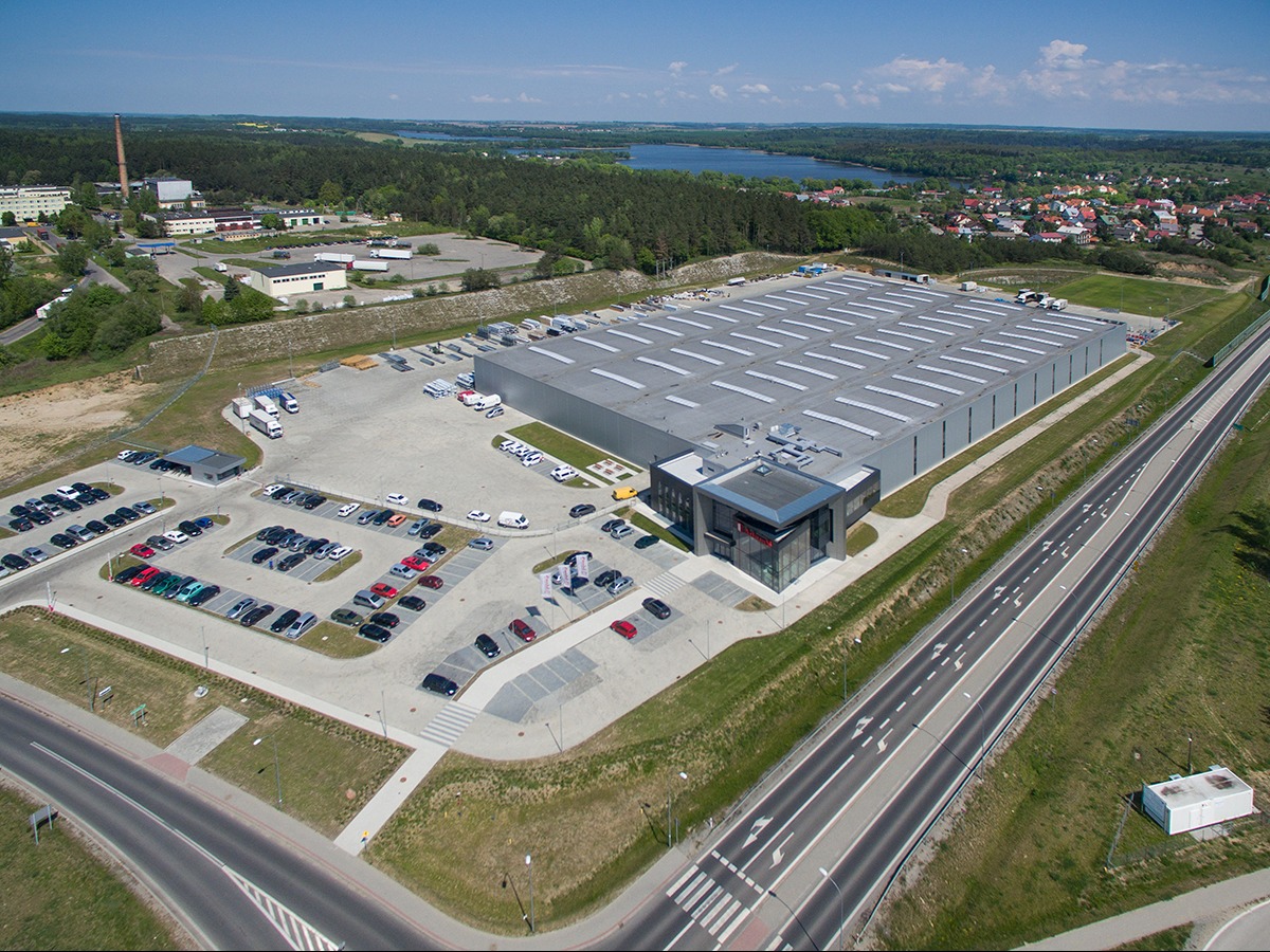 Widok z lotu ptaka na nowoczesny budynek magazynowo-biurowy z dużą ilością świetlików dachowych, przyległym parkingiem i drogą dojazdową, w tle las i zabudowa miejska.