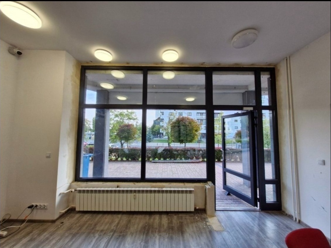 Wnętrze pomieszczenia z dużą, przeszkloną ścianą z czarnymi profilami, otwartymi szklanymi drzwiami, grzejnikiem pod oknem i widokiem na ulicę z zielenią.